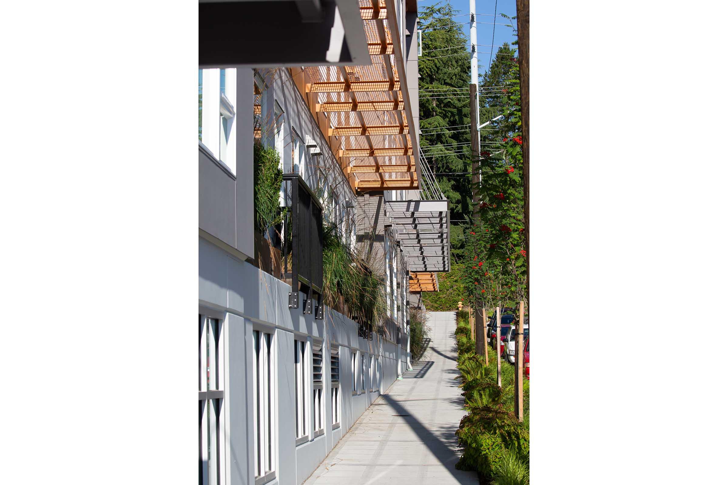 Modern building exterior featuring large windows and attractive architectural elements. A sidewalk runs alongside the structure, lined with greenery and trees. The upper level has wooden awnings, and the overall design is clean and inviting, showcasing a combination of urban and natural elements.