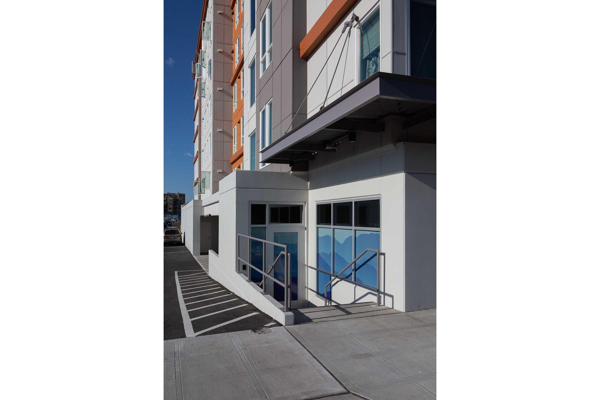 A modern building exterior featuring a white wall with large windows and an orange accent. The entrance has a ramp with railings for accessibility. The sidewalk is neatly paved, and there are parking spaces visible in the background under a clear blue sky.