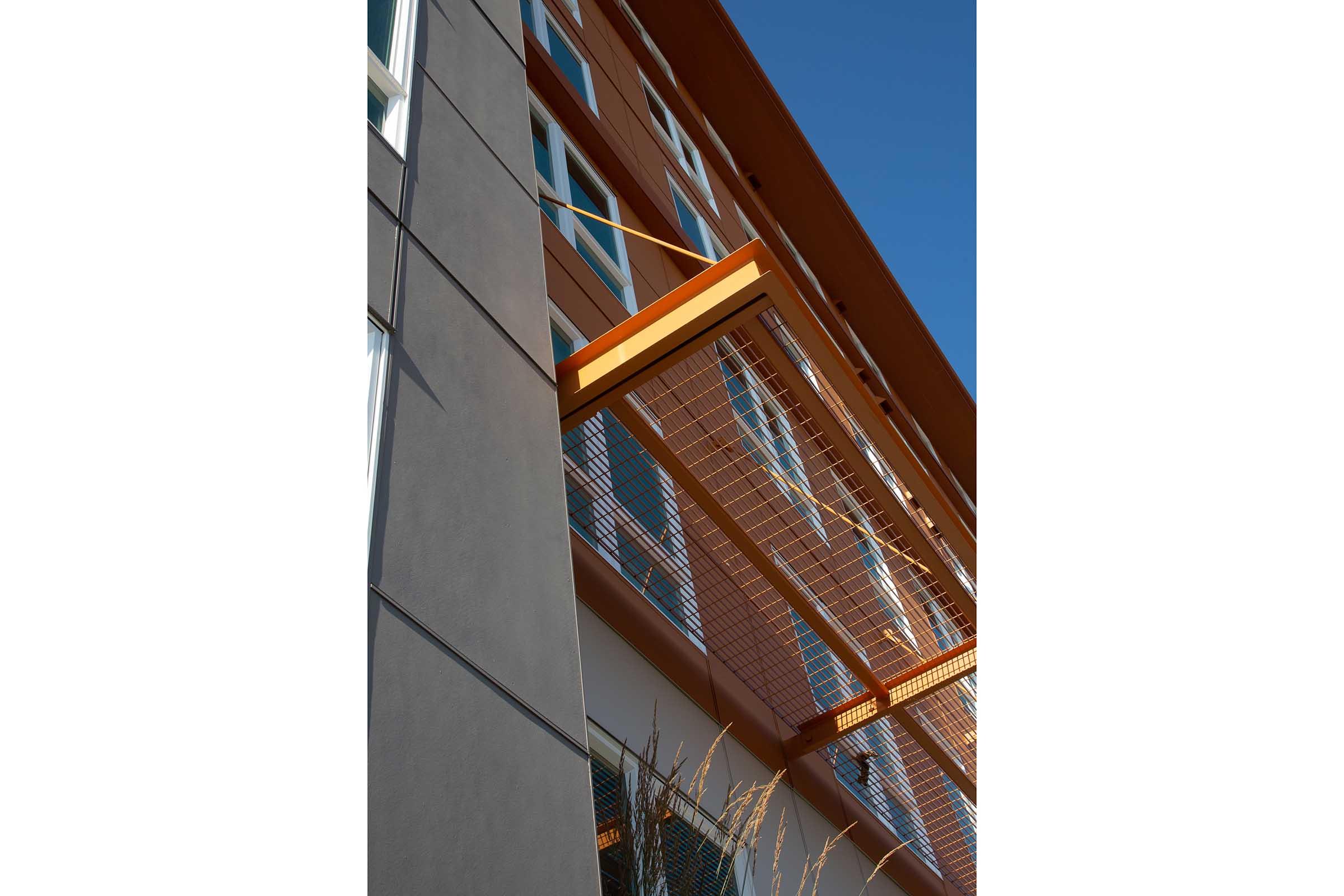 A modern building featuring a combination of textured gray and warm brown panels. A prominent orange metal awning extends above a row of large windows, with a clear blue sky in the background. The structure has a clean and contemporary architectural style.