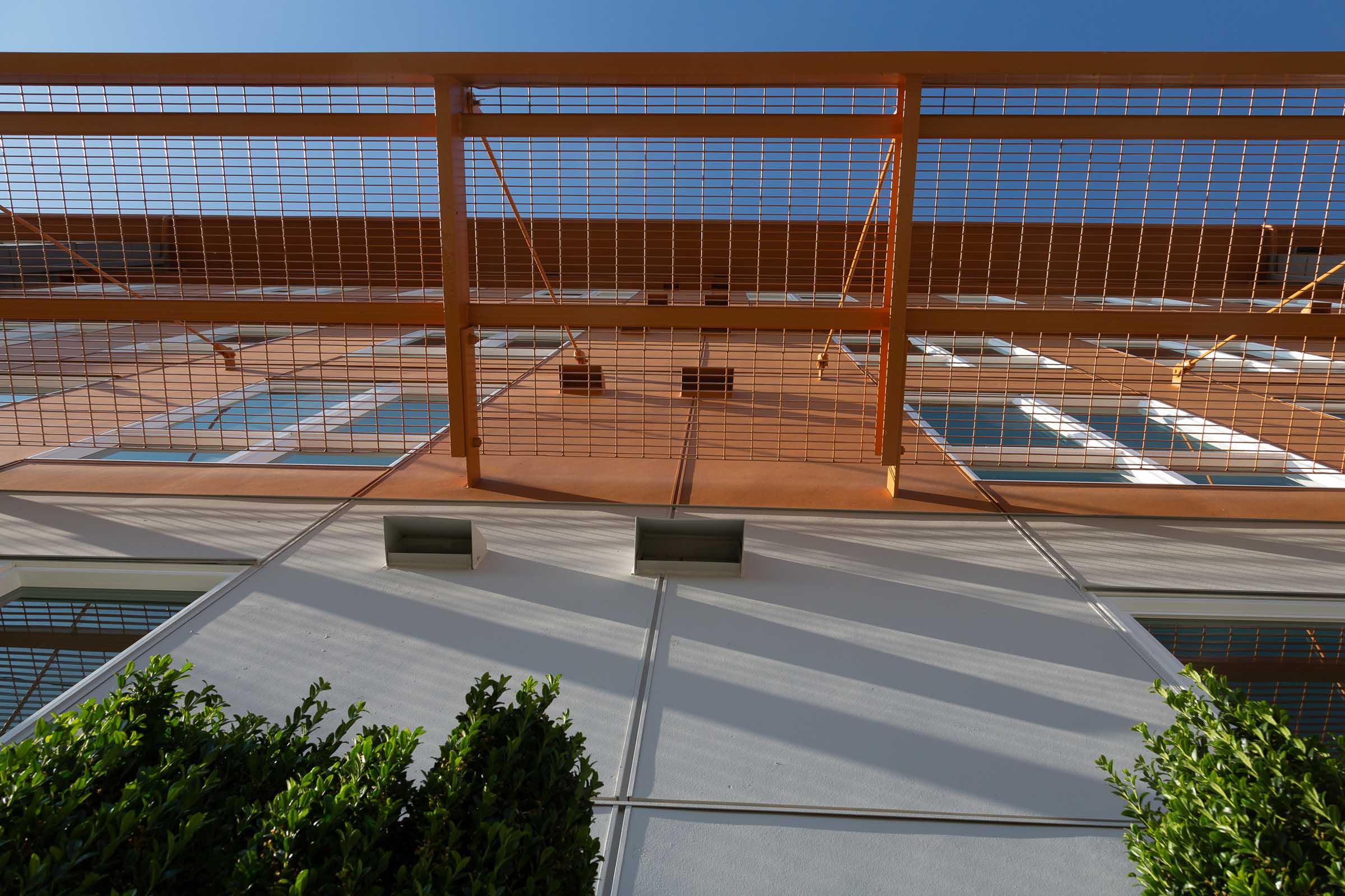 A low-angle view of a modern building with a textured orange facade and multiple windows. A metal grid railing is visible above, and neatly trimmed green bushes line the base of the structure. The clear blue sky provides a bright backdrop, enhancing the architectural features.