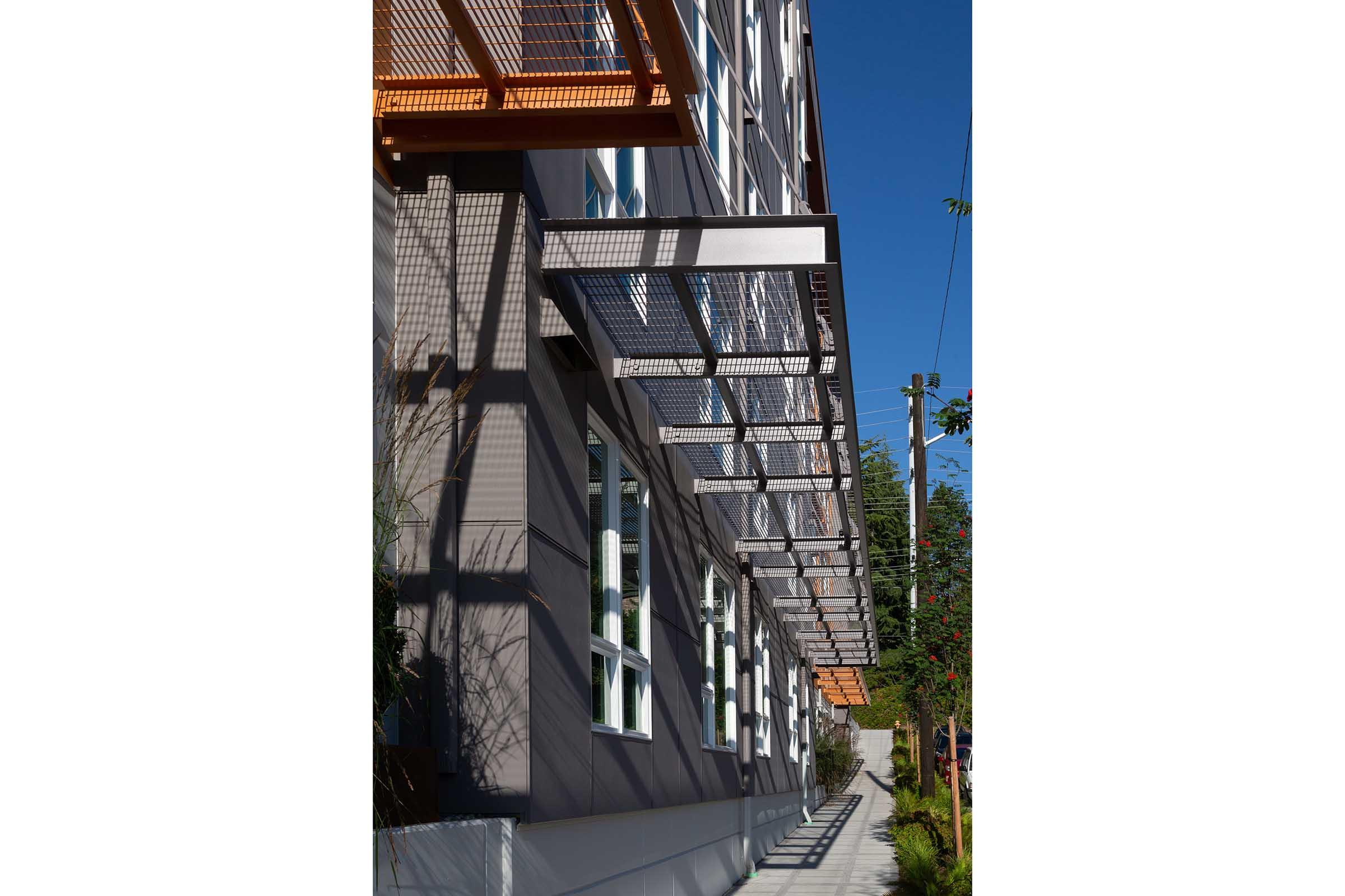 A modern building exterior featuring large windows with metal awnings, casting intricate shadows on the sidewalk. The facade includes geometric patterns and greenery, under a clear blue sky. The design reflects a contemporary architectural style.