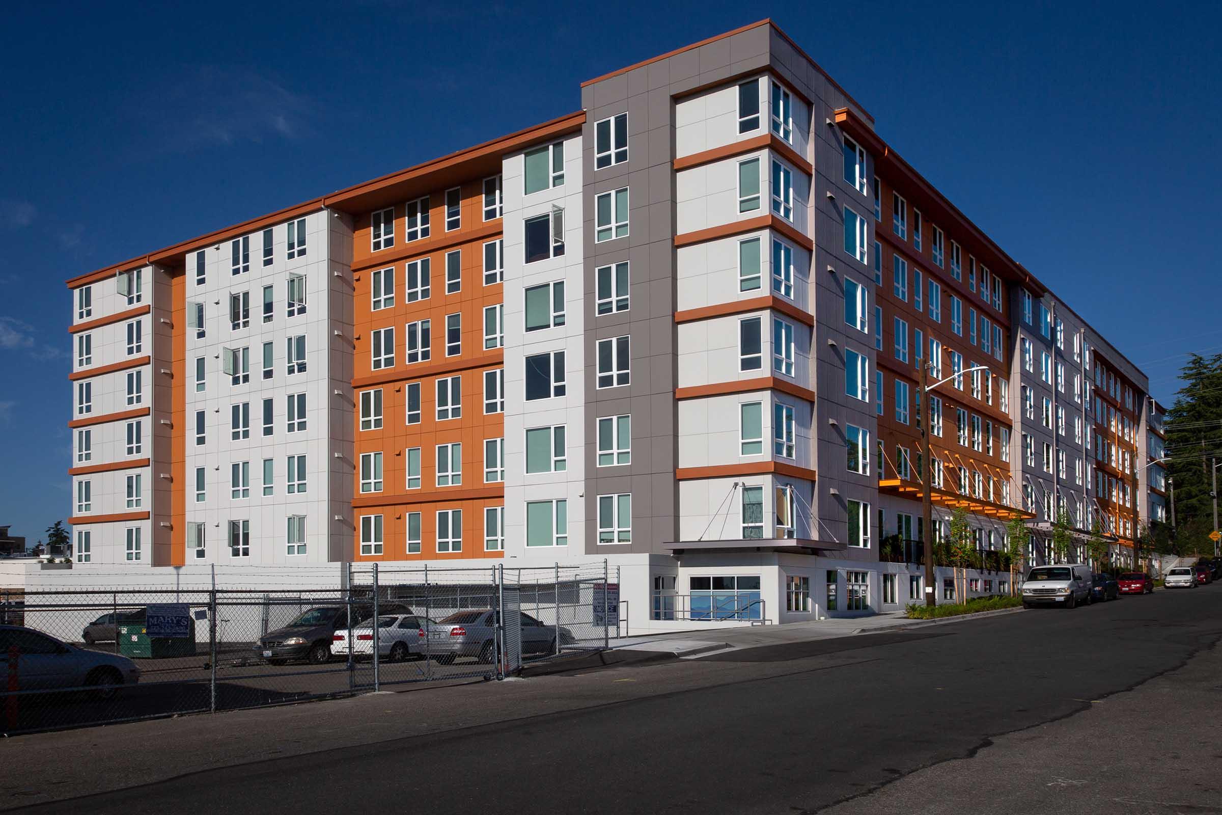 A modern multi-story apartment building with a mix of orange and white exterior panels. The building features numerous windows and balconies, set against a clear blue sky. In the foreground, there is a gravel parking area with several cars parked along the street, bordered by a chain-link fence.