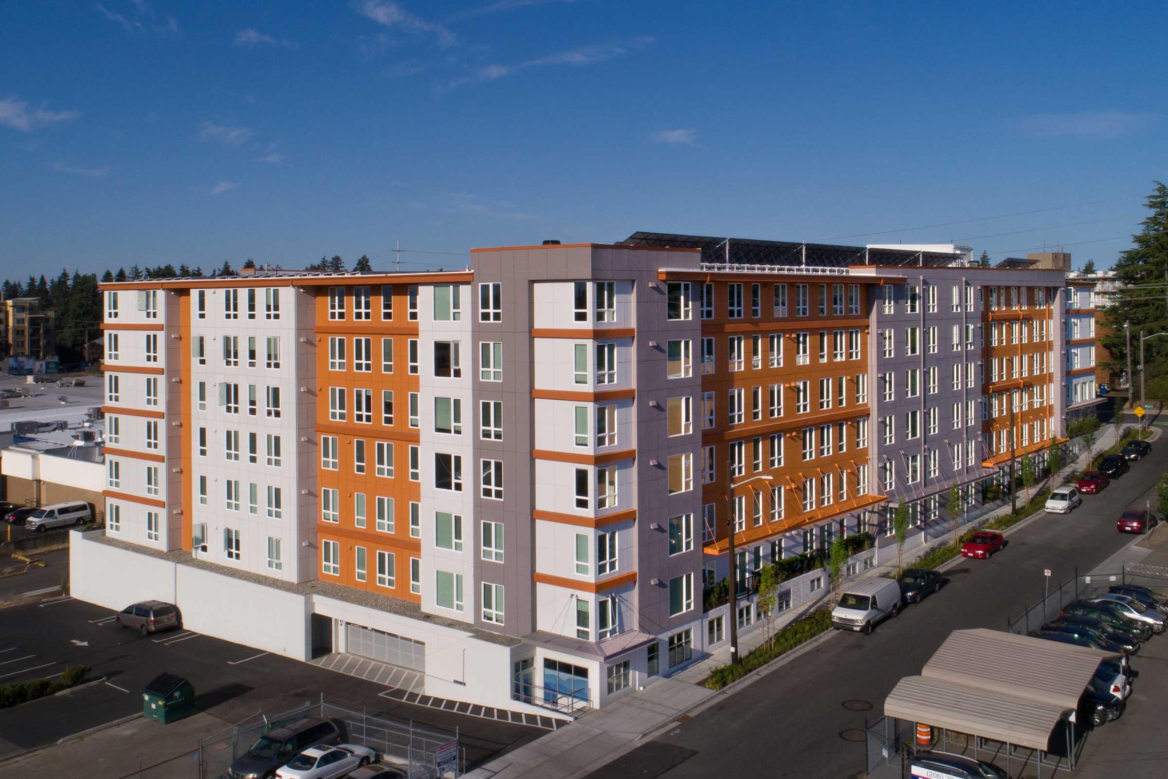 Modern multi-story apartment building with a mix of orange, gray, and white exterior. The structure features multiple windows and balconies, surrounded by a parking lot and street. Clear blue sky overhead, adding to the vibrant appearance of the urban landscape.