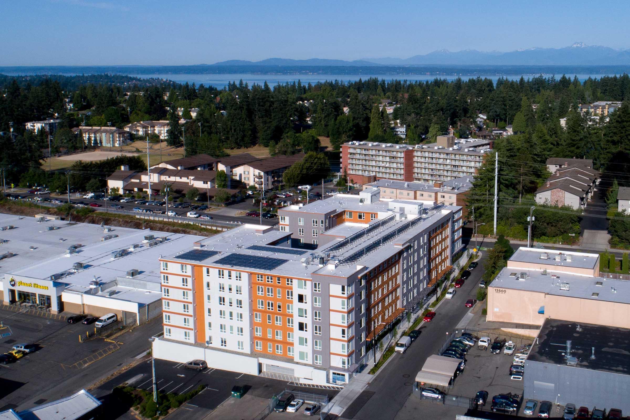 An aerial view of a multi-story building with a mix of orange, gray, and white exterior, surrounded by other residential and commercial structures. In the background, a scenic view of a lake and mountains under a clear blue sky. Parking lots and greenery are visible in the foreground.