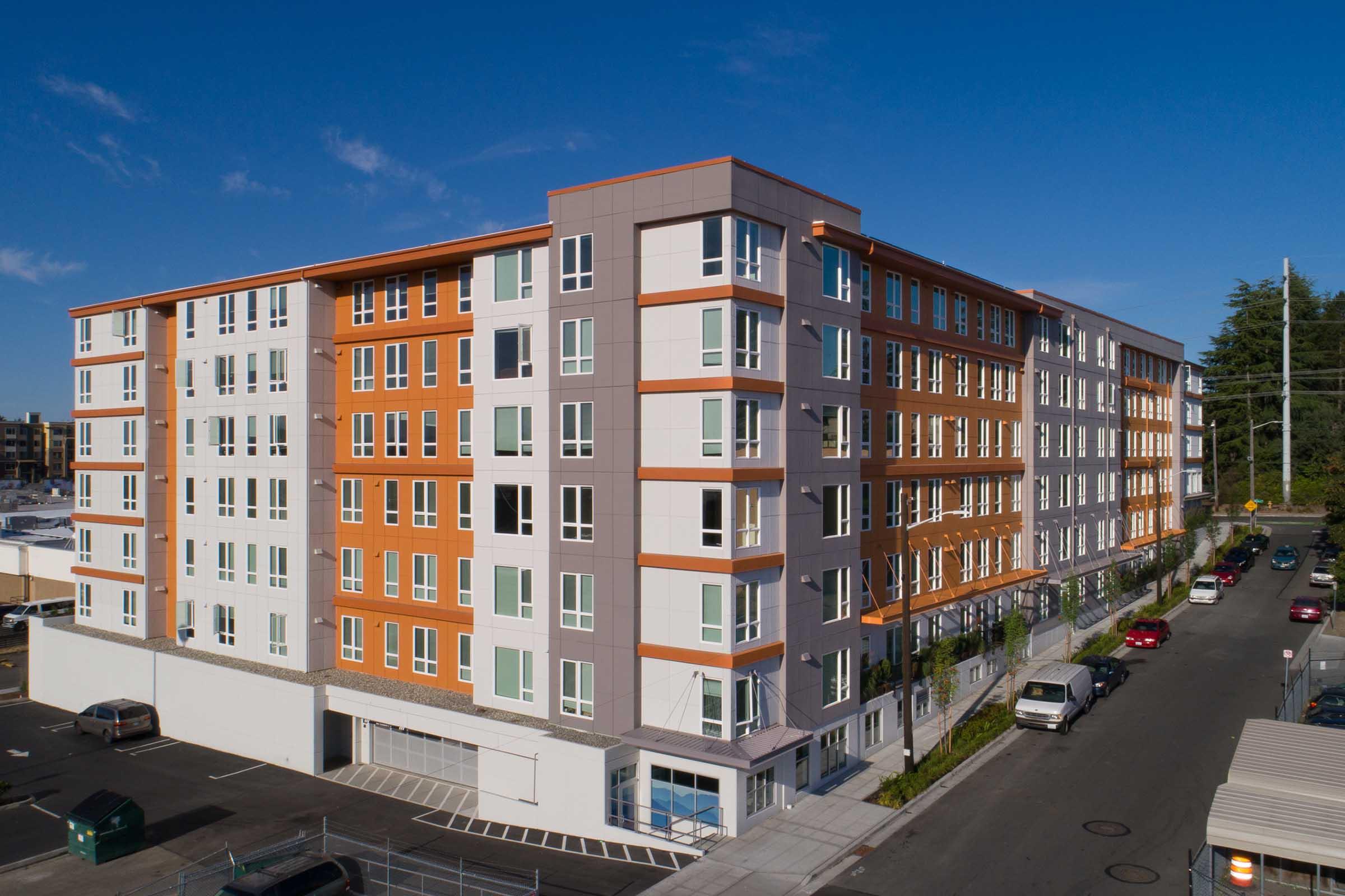 A modern residential building featuring a combination of white and orange exterior panels, with multiple floors of windows. The structure is situated on a city street, surrounded by parked cars and greenery. Clear blue sky above enhances the urban setting.