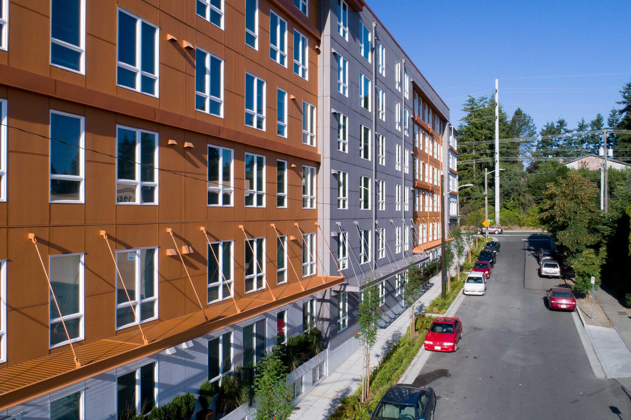 A modern residential building featuring a mix of brown and gray facades, large windows, and a landscaped walkway. The scene includes a street lined with parked cars and trees along the sidewalk, under a clear blue sky.