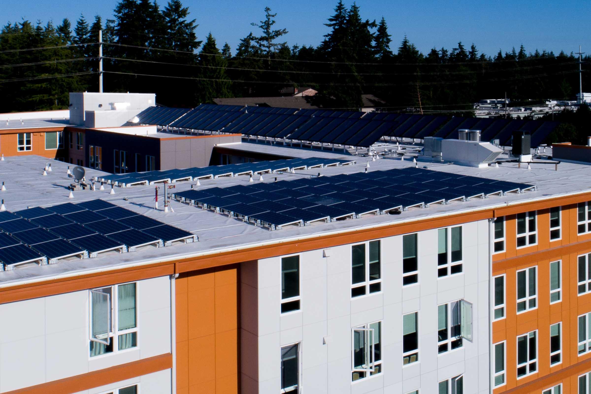 Aerial view of a modern building featuring a flat roof equipped with multiple solar panels. The building has a mix of orange and white exterior walls, surrounded by trees and blue sky. Solar panels are prominently installed on the roof, indicating a focus on renewable energy.