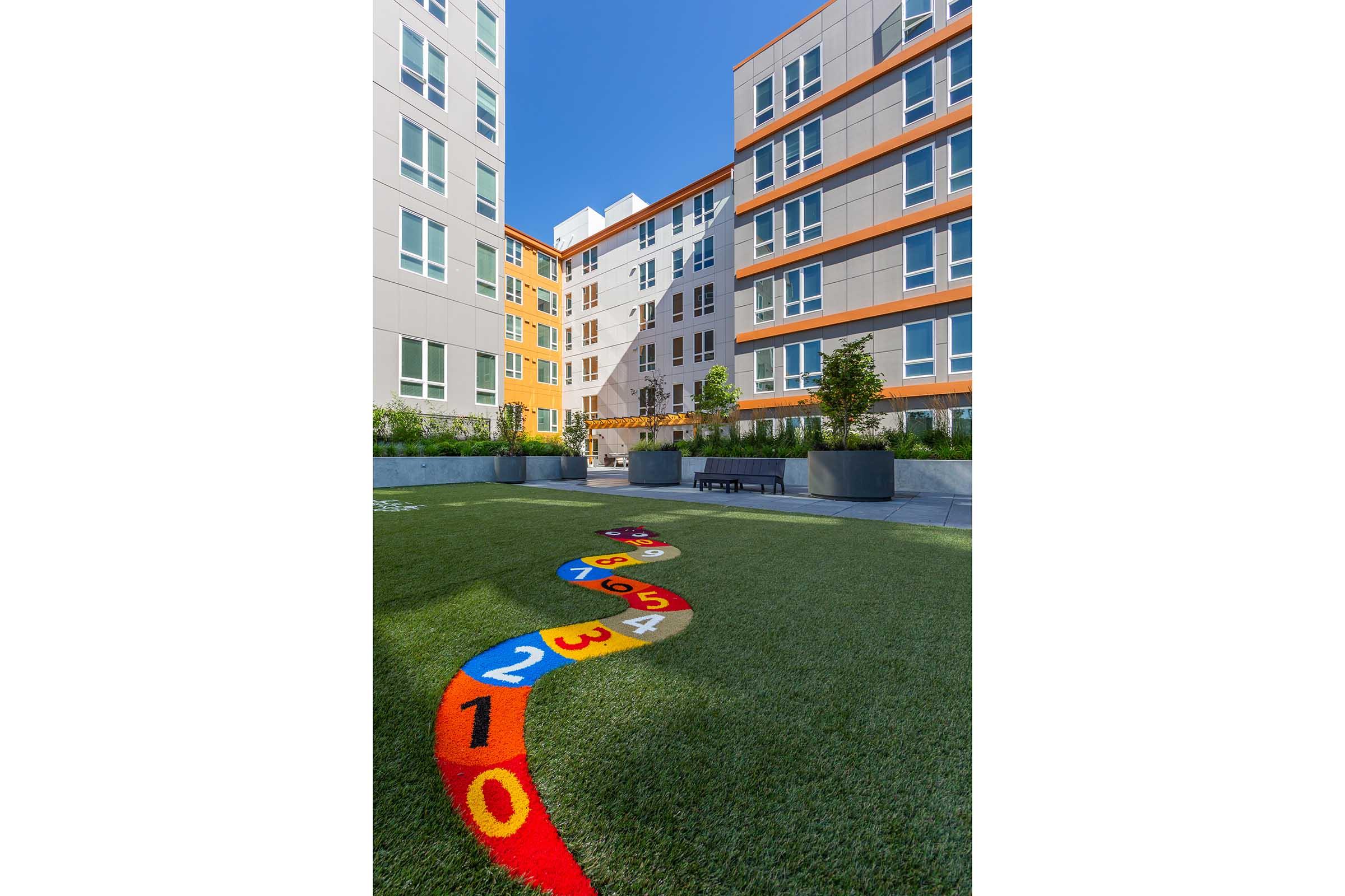 Colorful hopscotch game painted on artificial grass in a courtyard surrounded by modern residential buildings. The buildings feature a mix of gray and vibrant orange exteriors, with small trees and planters enhancing the outdoor space. Clear blue sky above adds a bright atmosphere.