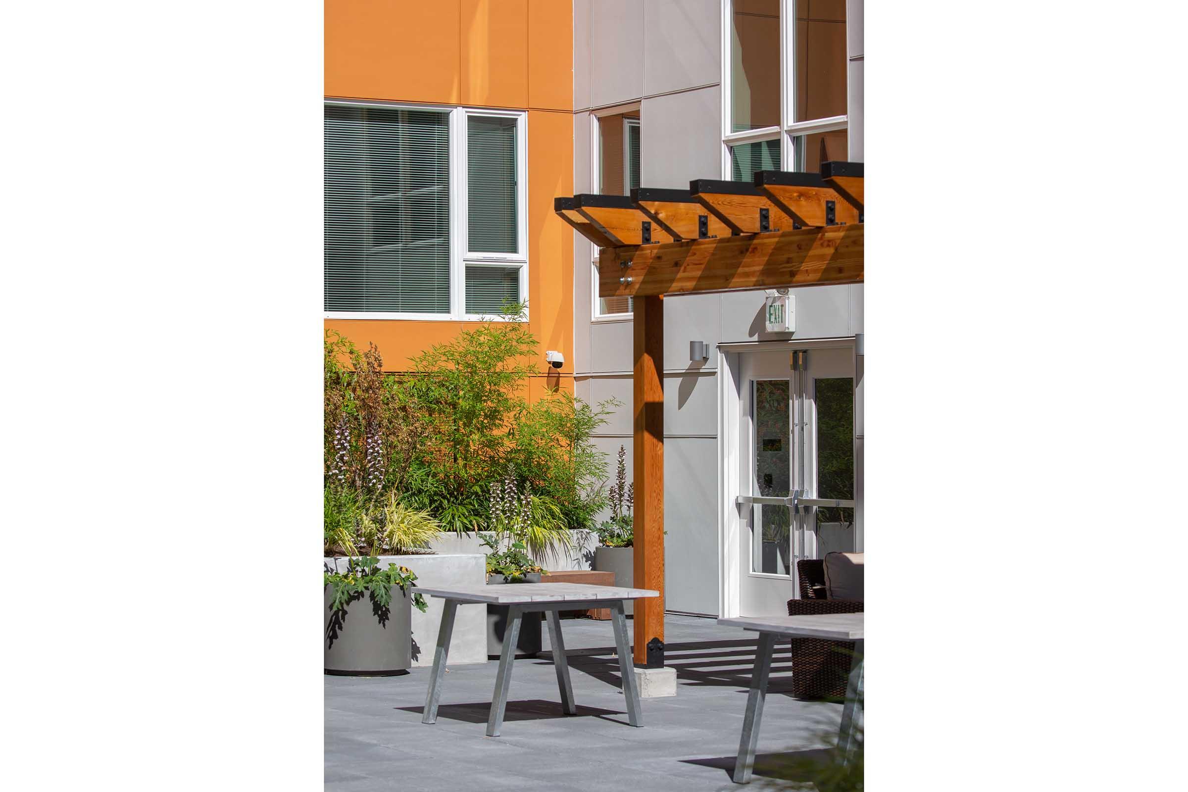 A modern outdoor patio area featuring a light wooden pergola, a gray picnic table, and lush green plants. The backdrop includes an orange-colored building with large windows, creating a vibrant and inviting atmosphere. Sunlight casts shadows on the patio floor, enhancing the serene setting.