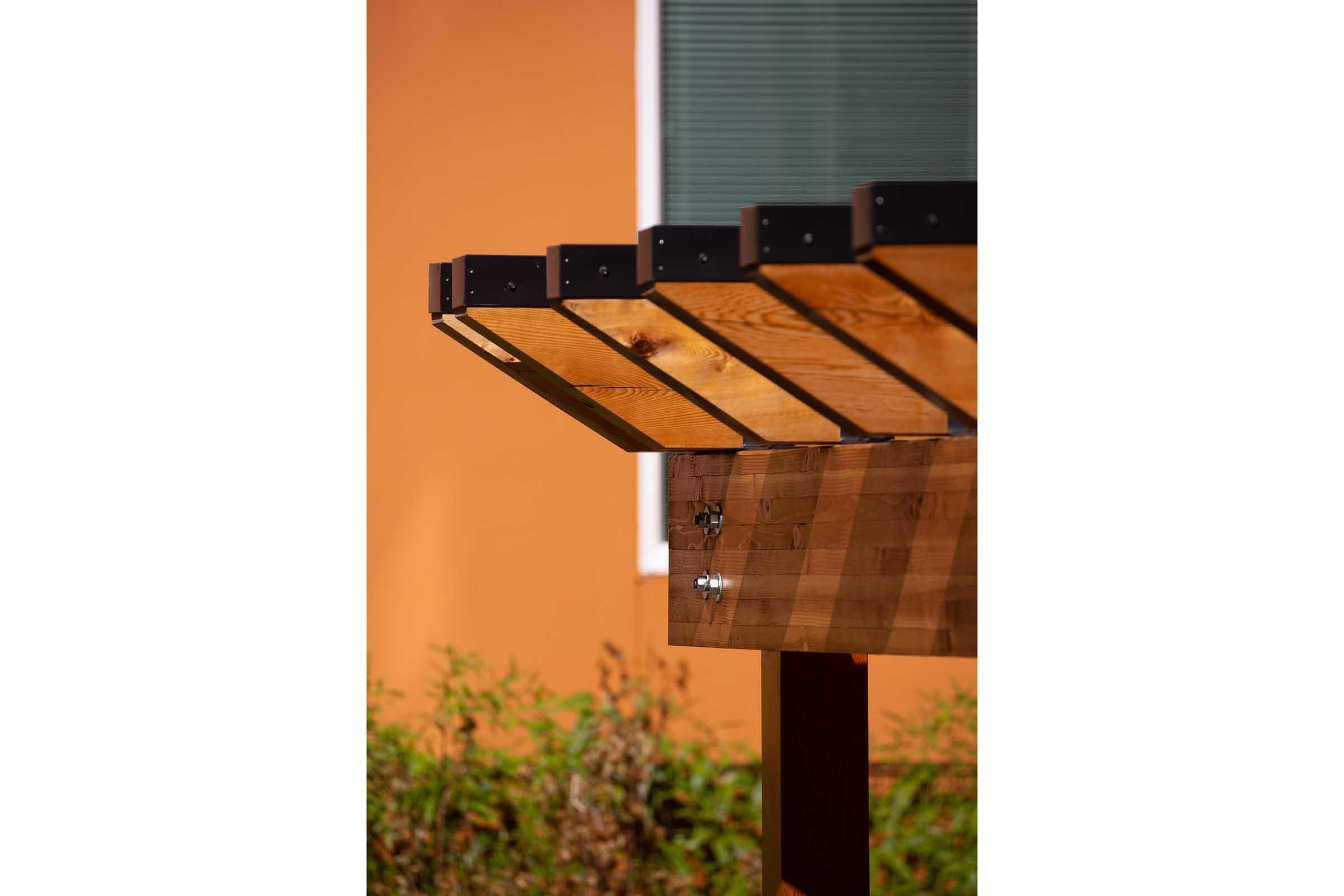 A close-up view of a wooden pergola's slatted roof, showcasing the natural grain of the wood and metal fasteners. In the background, there is an orange wall and some greenery, highlighting the structure's design and materials.