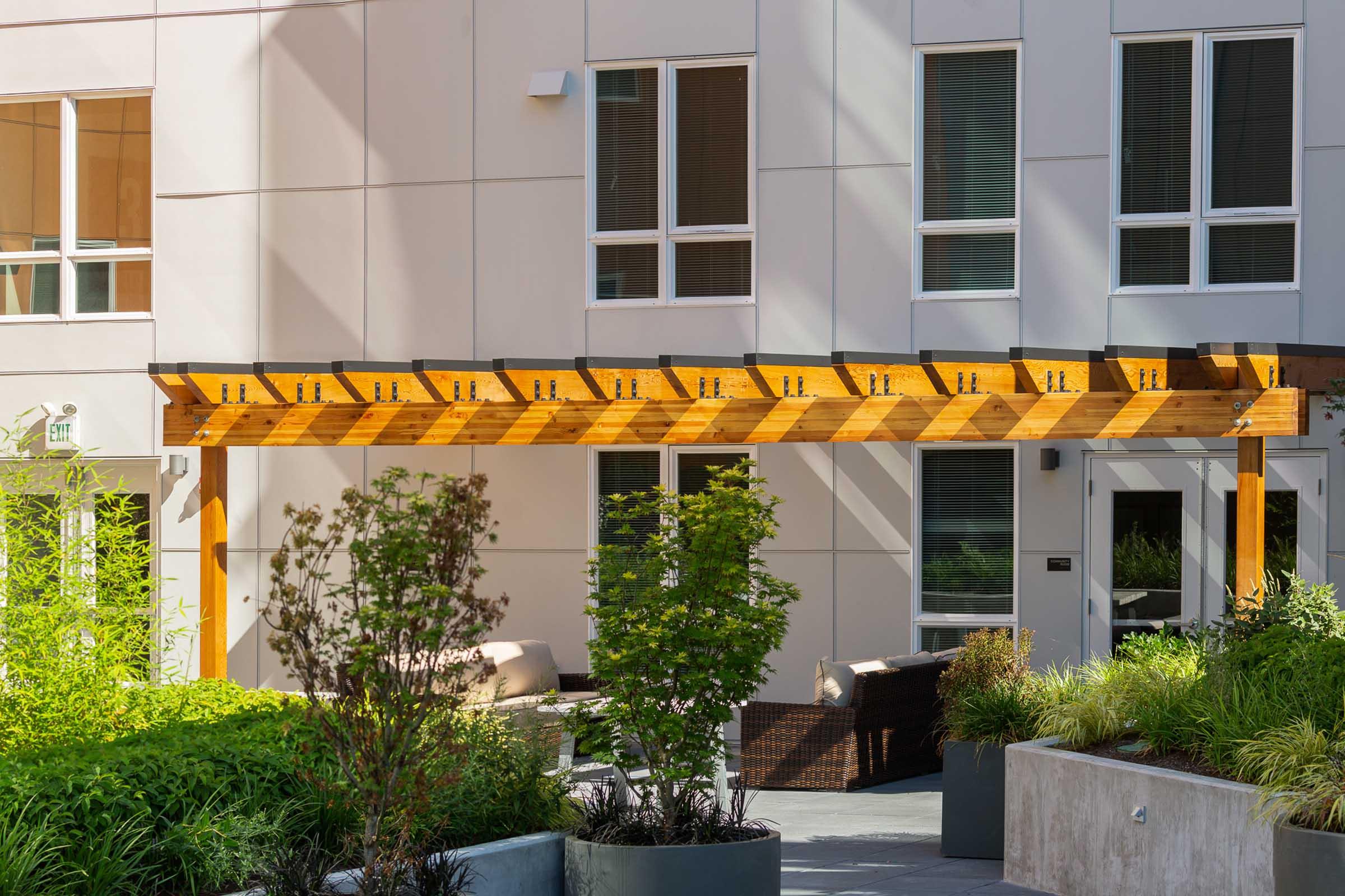 A wooden pergola with hooks for hanging items is situated in a landscaped outdoor area. The backdrop features a modern building with large windows, and there are green plants and shrubs in decorative planters around the space, creating a welcoming atmosphere.