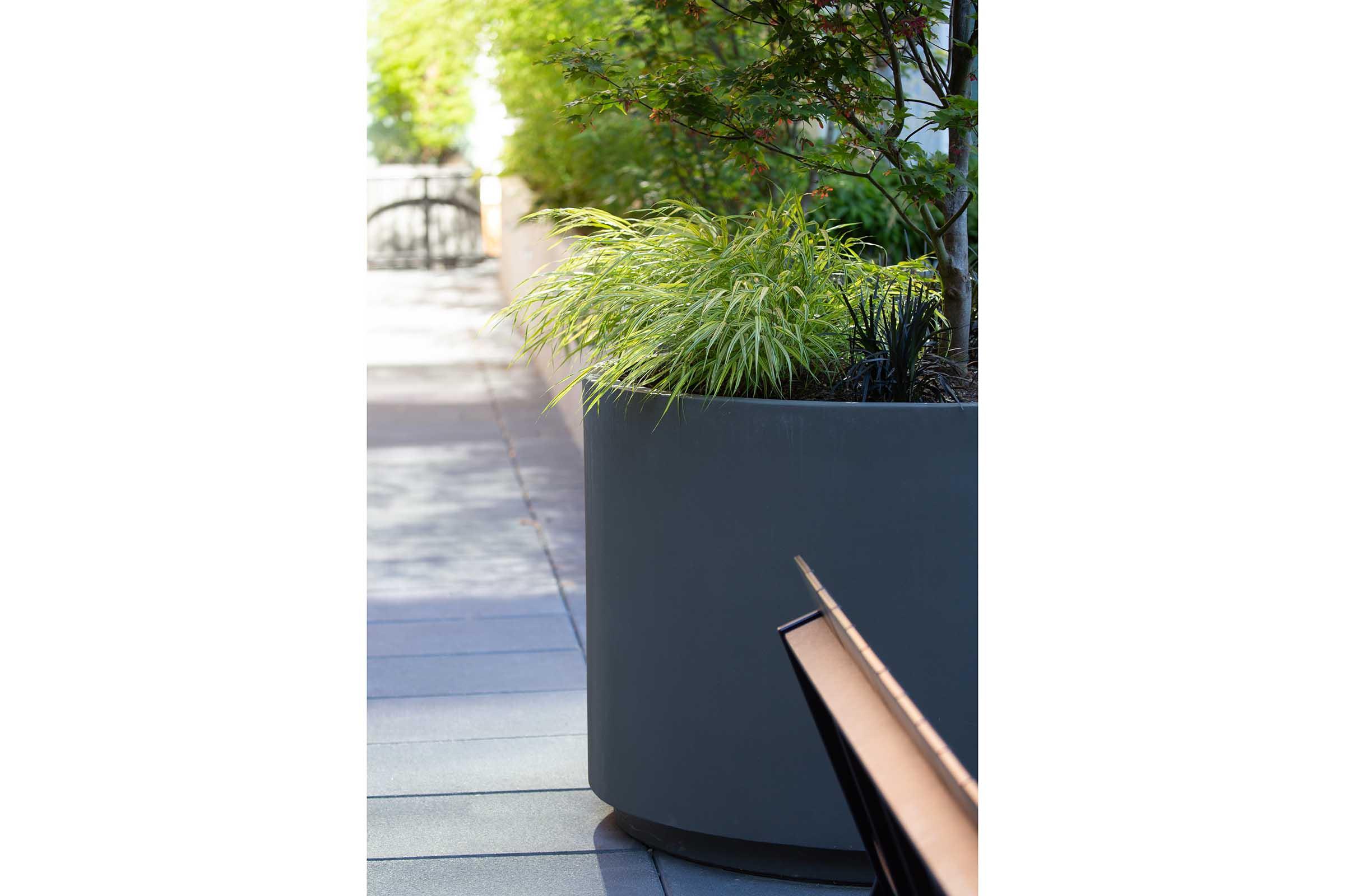 A modern outdoor setting featuring a large, round gray planter filled with lush green grass and plants. In the background, there is a blurred walkway and foliage, creating a serene atmosphere. A wooden bench is partially visible in the foreground, adding to the tranquil scene.