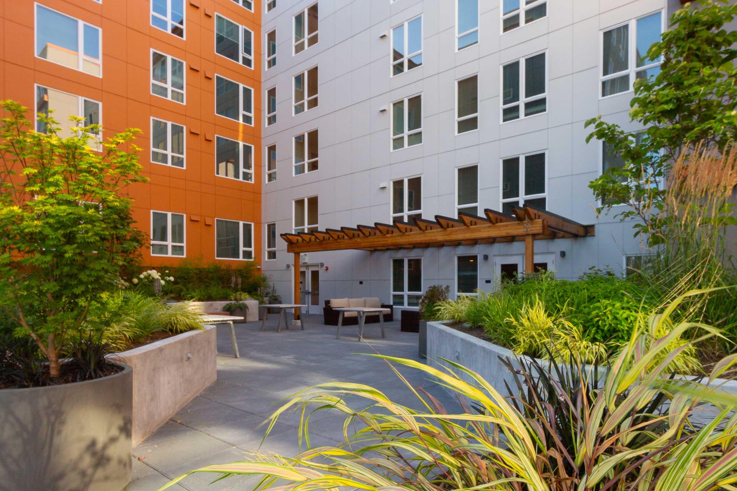 A modern courtyard surrounded by two multi-story buildings with orange and gray facades. The area features landscaped planters filled with greenery, a wooden pergola, and various seating options, creating a welcoming outdoor space for relaxation and gathering.