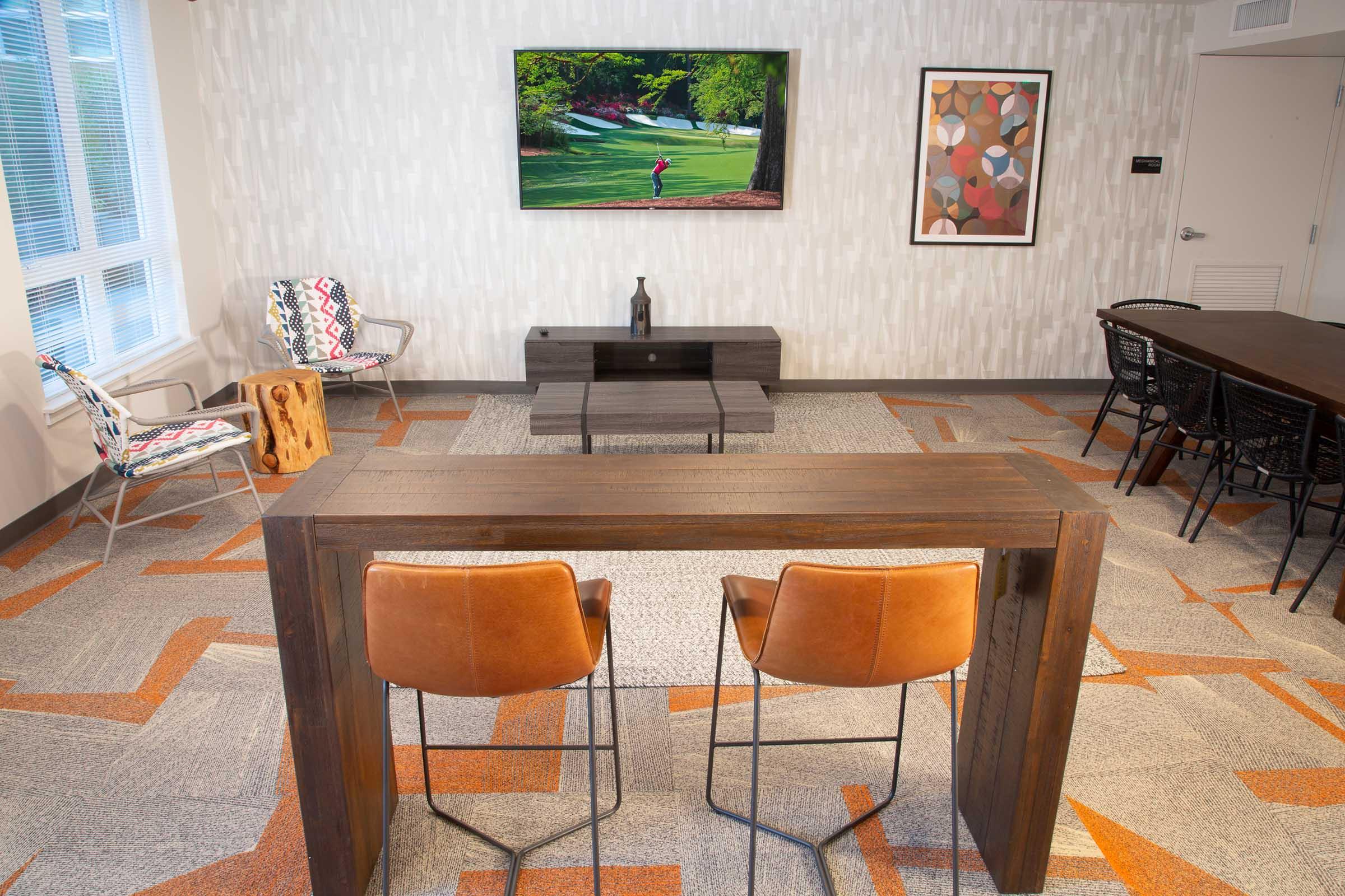 A modern, stylish common area featuring a wooden bar table with two brown leather stools, a sleek black coffee table, and a wall-mounted TV displaying a golf scene. The space is decorated with colorful artwork and has a cozy, inviting design, complemented by patterned carpet and bright natural light.