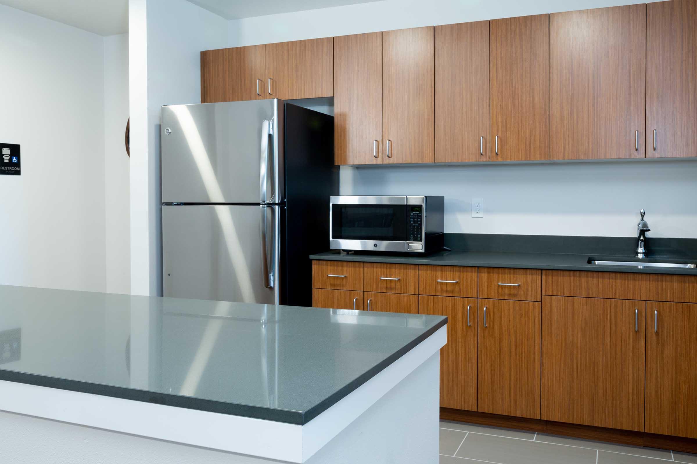 Modern kitchen featuring wooden cabinetry, a stainless steel refrigerator, and a microwave on a black countertop. The spacious layout includes an island with a gray surface, and minimalist design elements create a clean and contemporary look. The walls are painted white, enhancing the bright atmosphere.