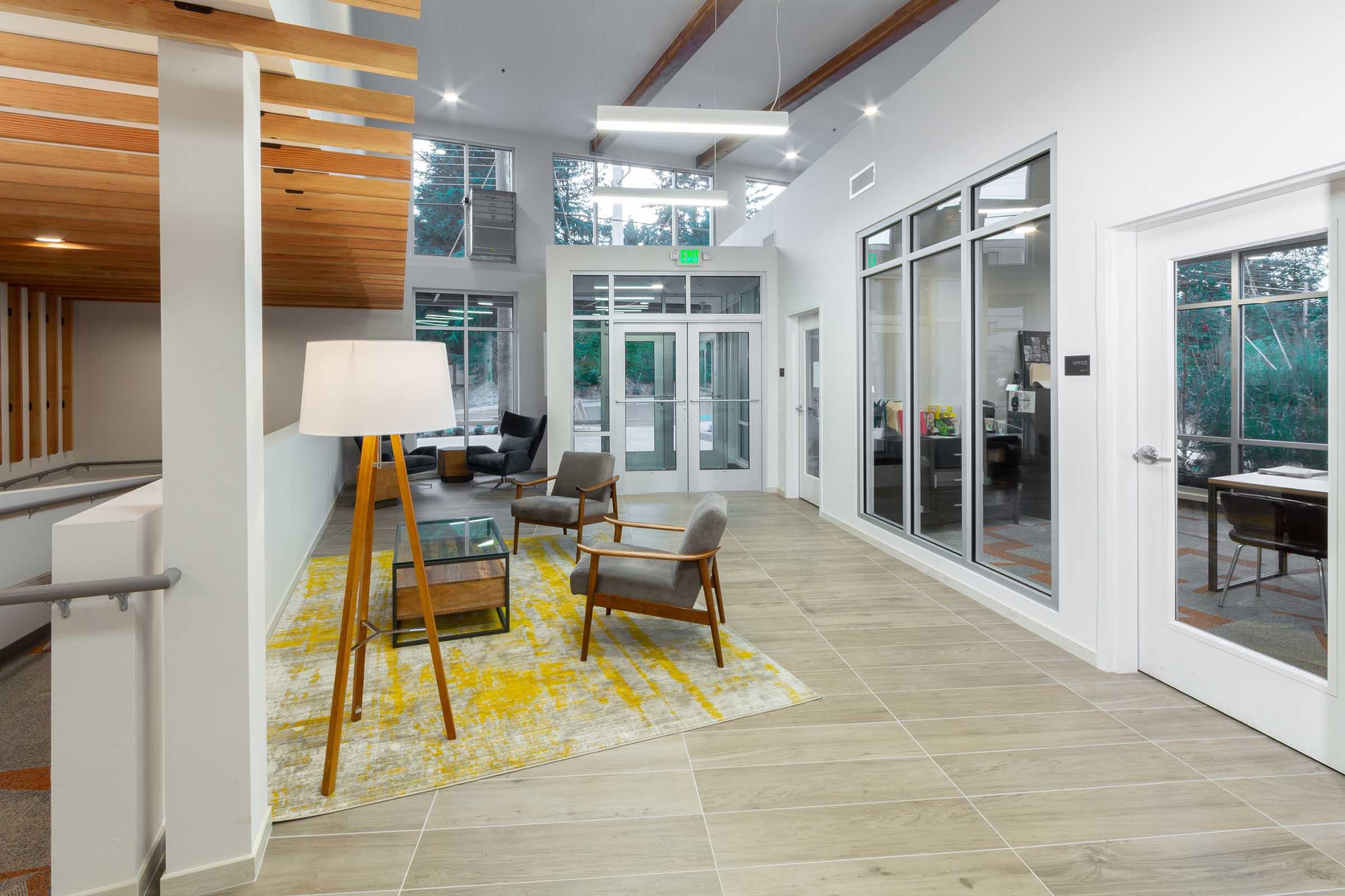 A modern interior space featuring a bright, open layout with large windows. There are two gray armchairs and a wooden coffee table on a yellow-patterned rug. A standing lamp is in the corner, and glass doors lead to an outdoor area. The ceiling has wooden beams, and the floor is tiled in light colors.