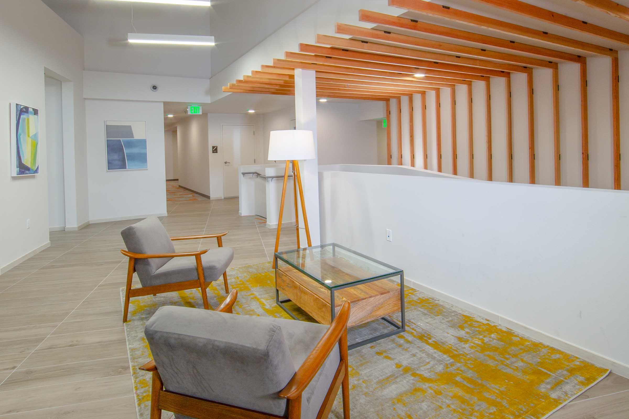 A modern lounge area featuring two stylish armchairs, a glass-top coffee table, and a decorative rug. The space has a clean, minimalist design with light-colored walls, wooden accents on the ceiling, and soft lighting, creating a welcoming atmosphere.