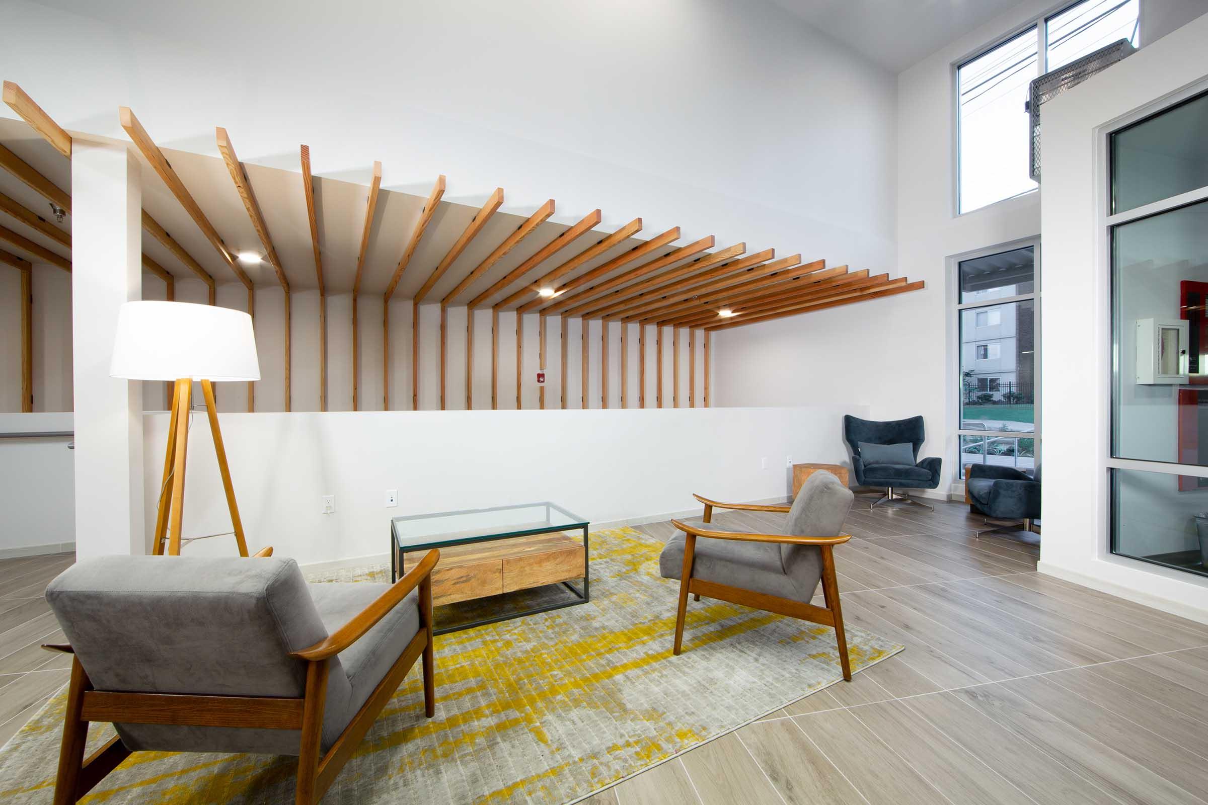 A modern interior space featuring a minimalist design with wooden slats on the wall, two comfortable mid-century style chairs, a glass coffee table on a yellow and gray patterned rug, and a floor lamp. Large windows allow natural light to fill the area.