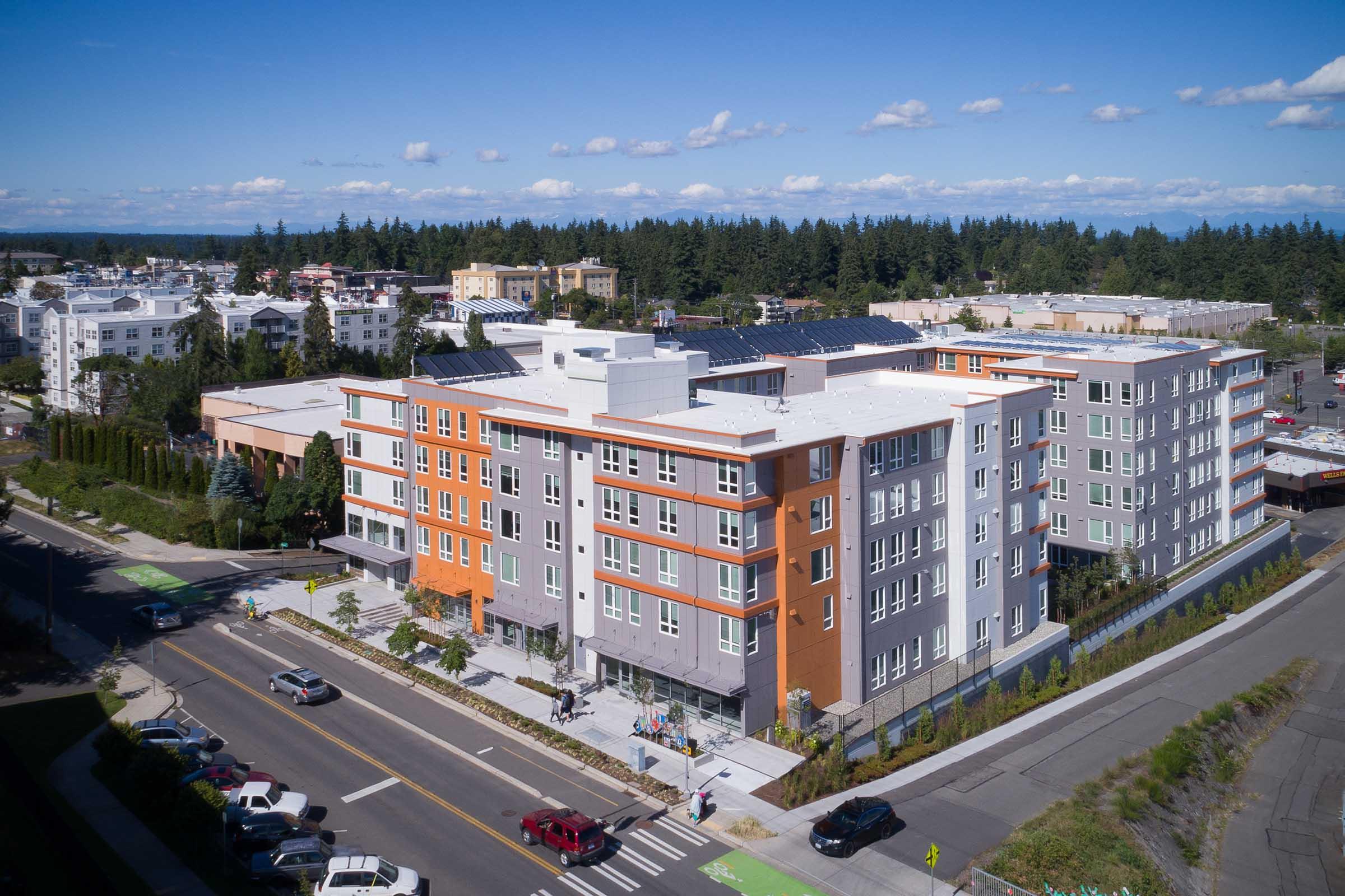 Aerial view of a modern multi-story apartment building featuring a mix of orange, gray, and white exteriors, surrounded by trees and a portion of an urban landscape. Streets with parked cars and pedestrians are visible in the foreground, indicating a vibrant community environment. Blue sky and clouds above.