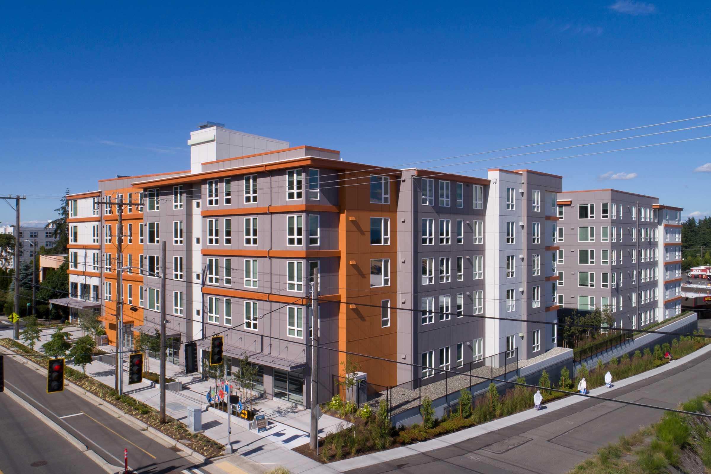 Modern multi-story residential building featuring a mix of orange and gray exterior panels, large windows, and landscaped surroundings. The structure is situated near a busy street with traffic lights and pedestrian access. Clear blue sky visible overhead.