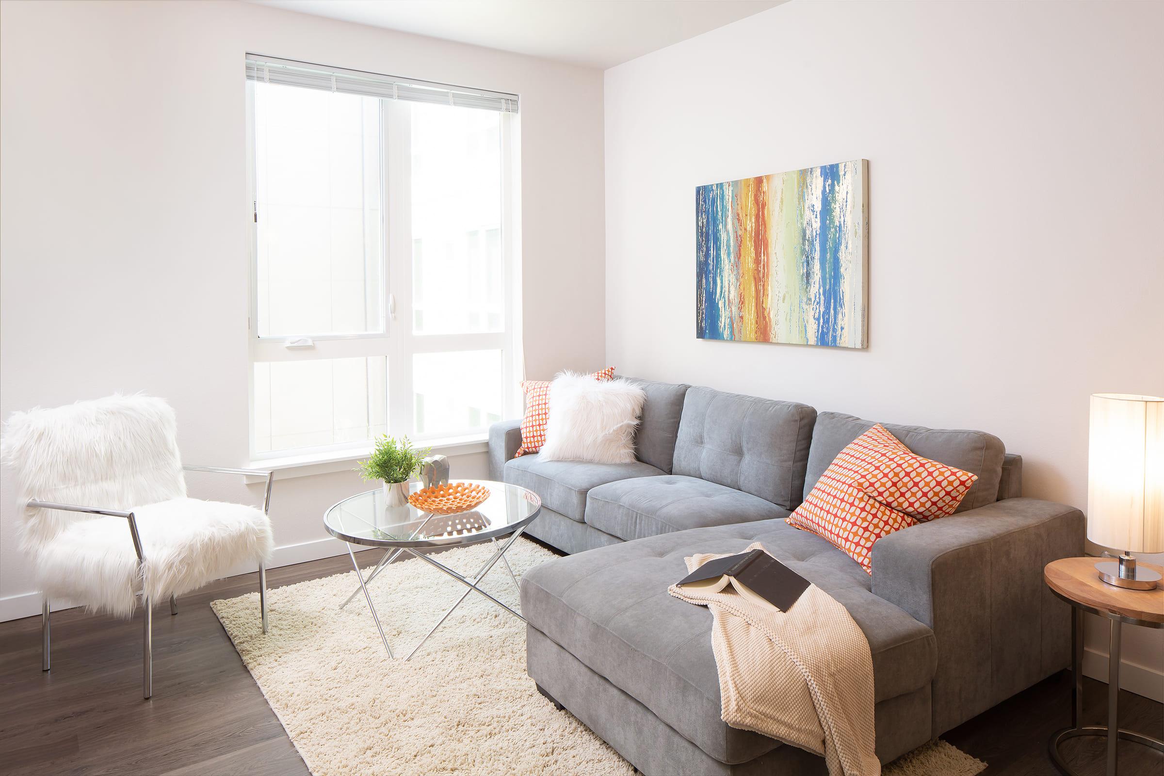 A modern living room featuring a gray sectional sofa with colorful throw pillows, a glass coffee table, and a cozy armchair. A fluffy white rug covers the dark wooden floor, and a piece of abstract wall art adds color. Natural light streams in through a large window.