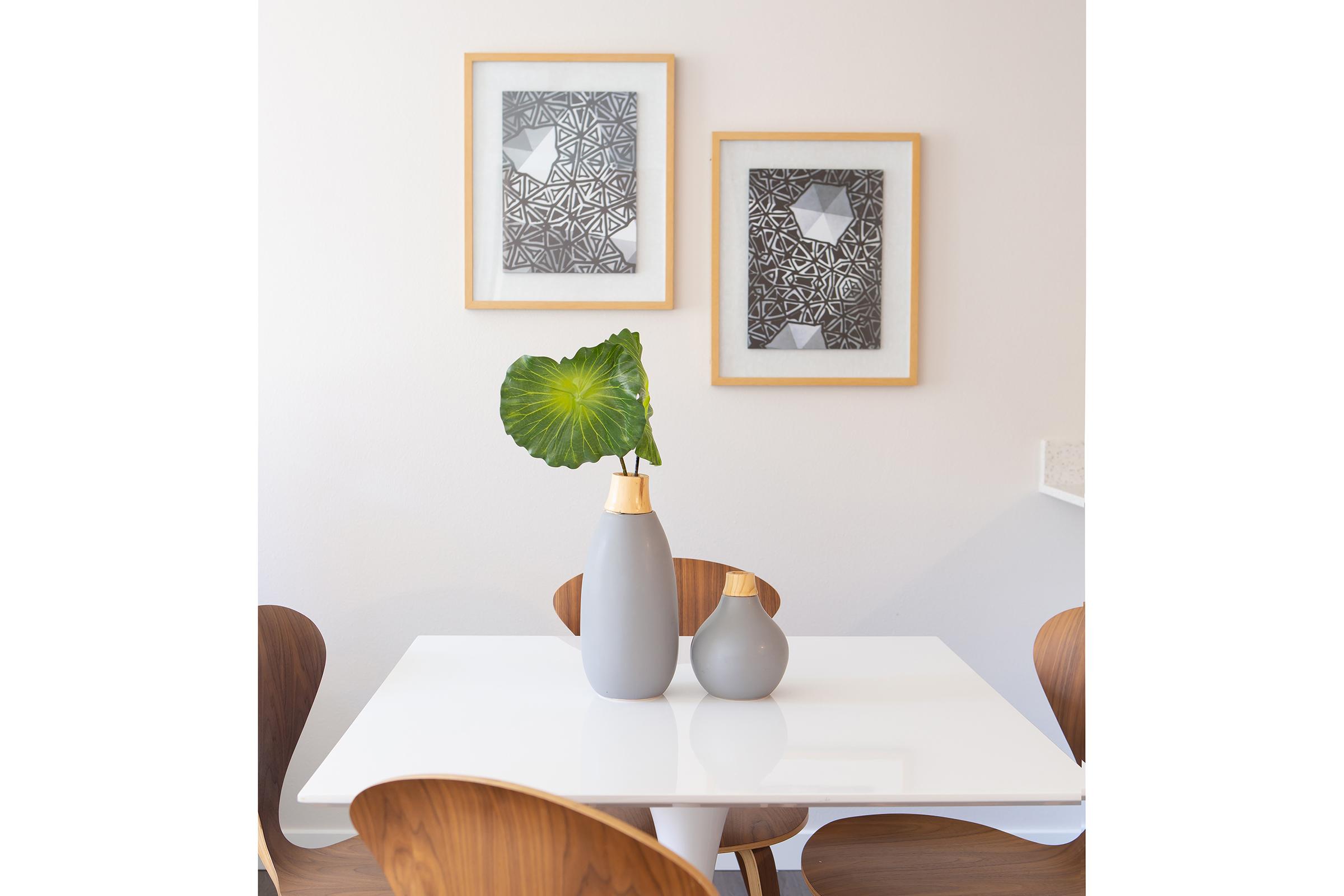 A modern dining area featuring a white table with two vases—one large and gray, the other smaller—holding a green leaf. Two framed abstract artworks in black and white hang on the wall behind. The chairs are wooden with a smooth, curved design, adding a contemporary touch to the space.