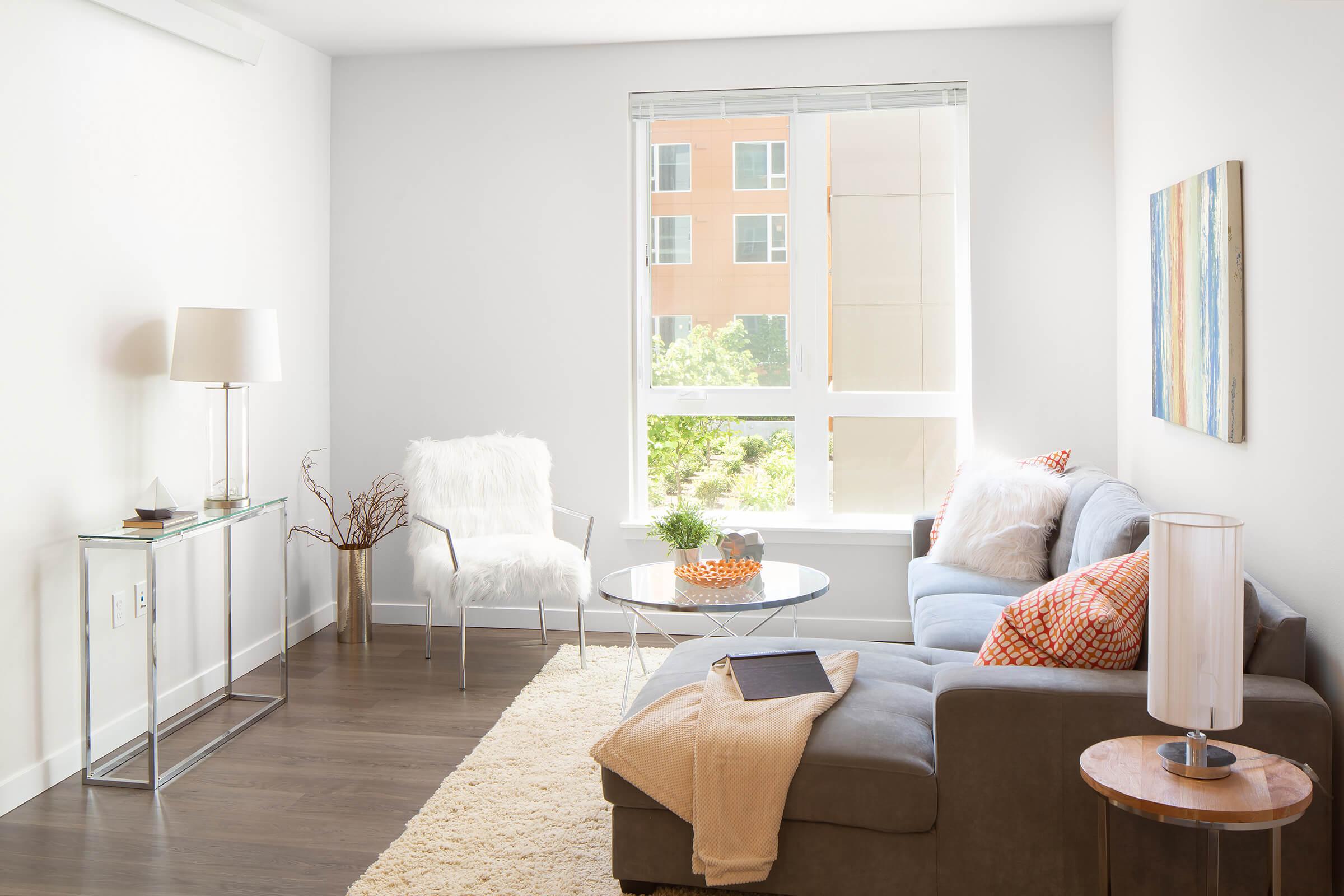 A cozy living room featuring a gray sofa with orange and white patterned cushions, a glass coffee table with a decorative bowl, a fluffy white chair, a side table with a lamp, and a light rug. Large windows allow natural light to fill the space, revealing greenery outside and modern architecture in the background.