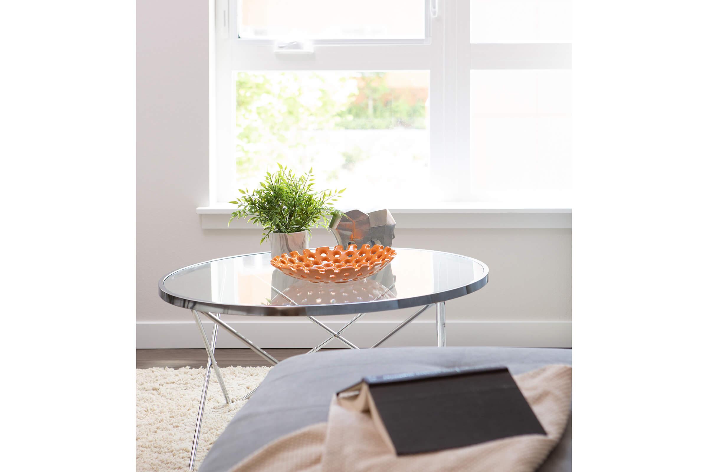 A modern living room scene featuring a round glass coffee table with a silver frame. On the table, there is a decorative orange bowl with a plant next to it. In the background, a large window lets in natural light, illuminating the soft textures of a light-colored rug and a cozy throw on the sofa.