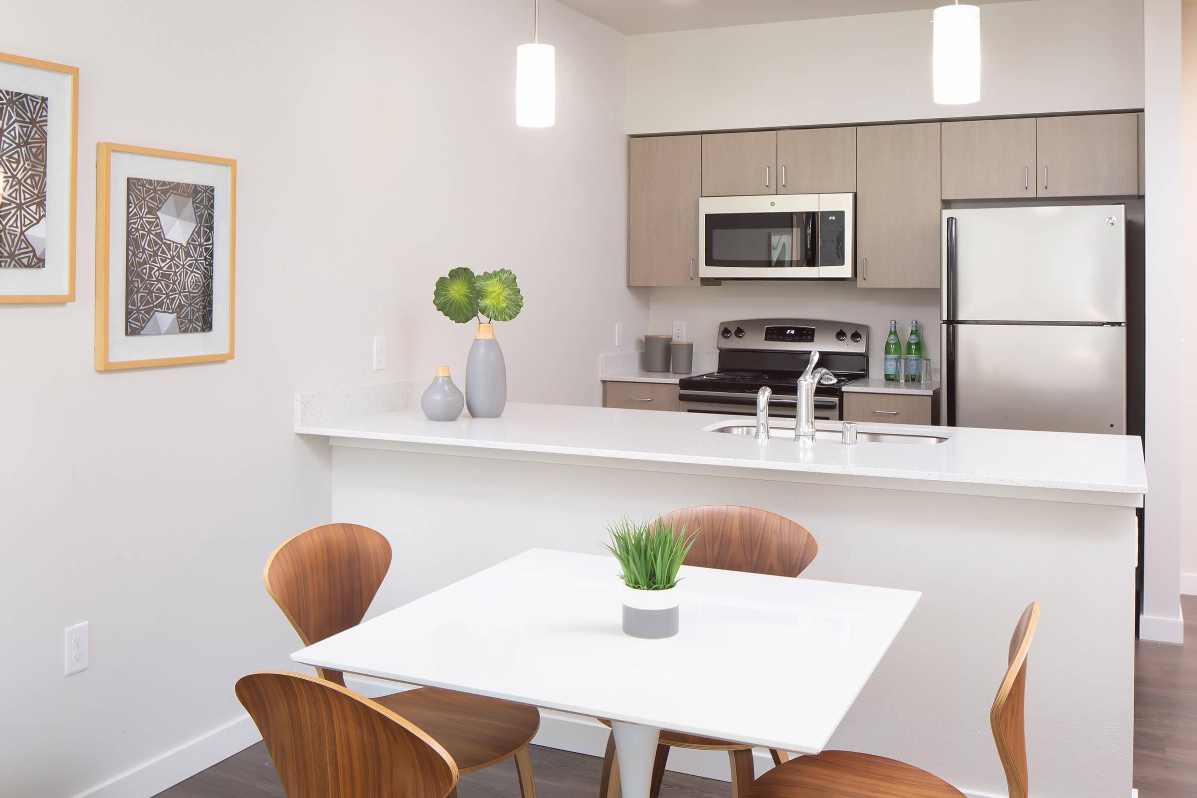 A modern kitchen with light wood cabinetry and stainless steel appliances, including a microwave and refrigerator. There is a white countertop with a small plant and decorative vases, along with a dining area featuring a white table and four wooden chairs. Two framed artworks are hung on the wall.