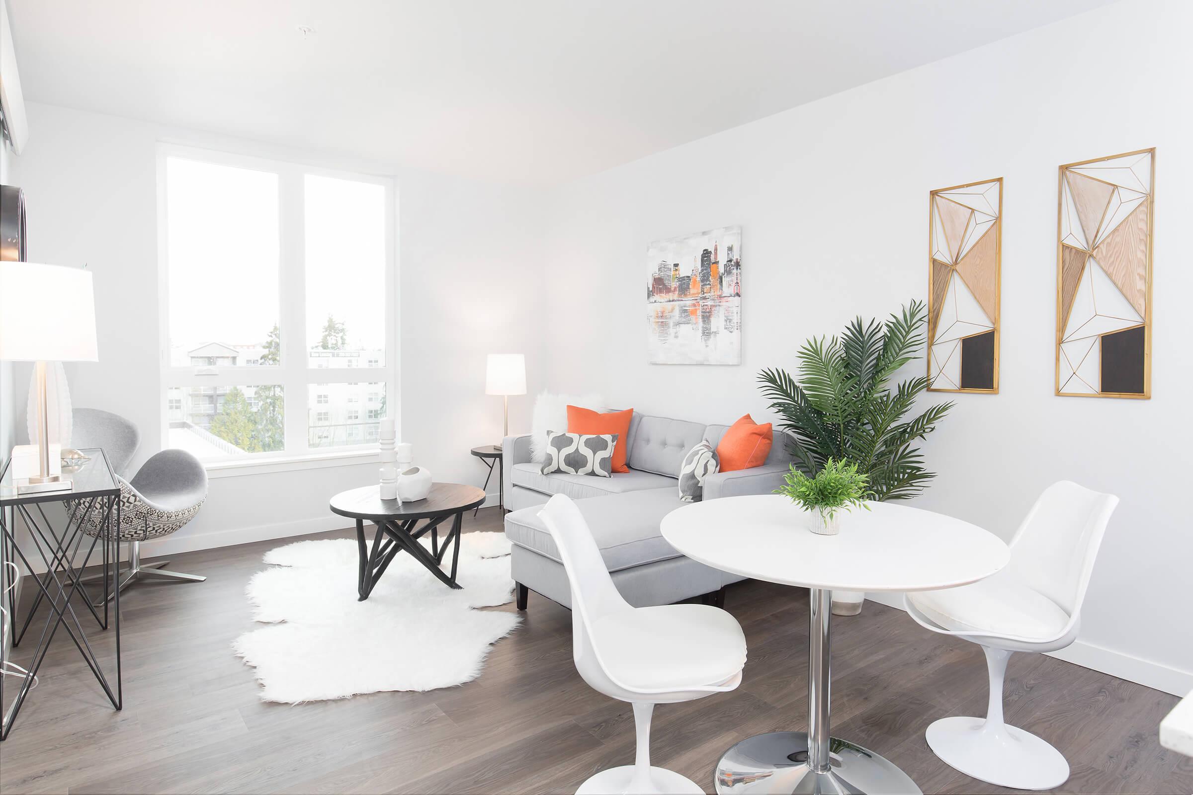 A modern living room featuring a gray sofa with orange accent pillows, a round coffee table on a white rug, and a small dining area with a white table and chairs. Two decorative wall hangings are visible, along with a small potted plant, a lamp, and a stylish chair, all in a bright, airy space with large windows.