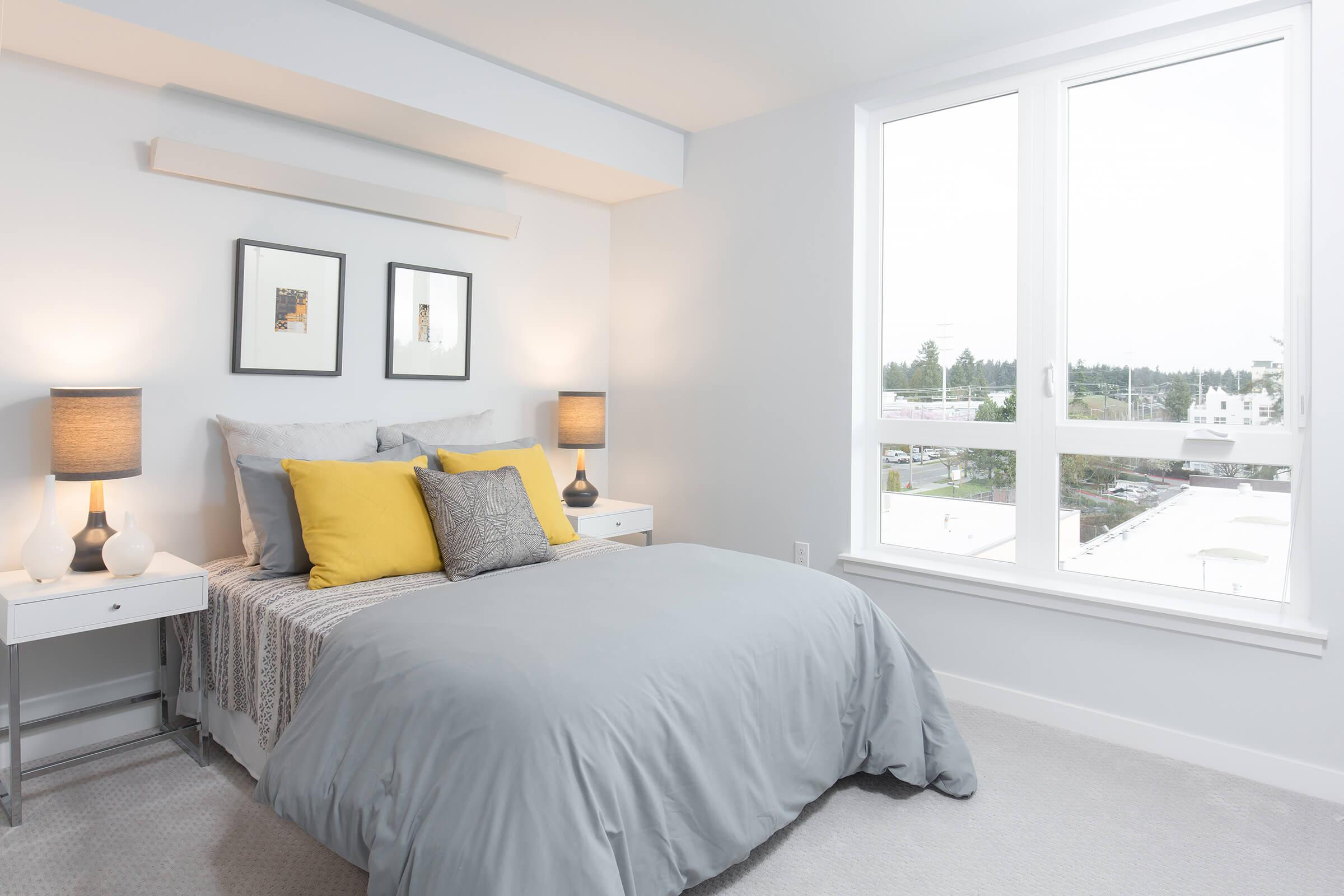 A cozy bedroom featuring a neatly made bed with gray bedding and yellow accent pillows. There are two bedside tables with lamps on either side of the bed, and two framed pictures on the wall. A large window lets in natural light, offering a view of the outdoors. Soft, neutral decor creates a calm atmosphere.