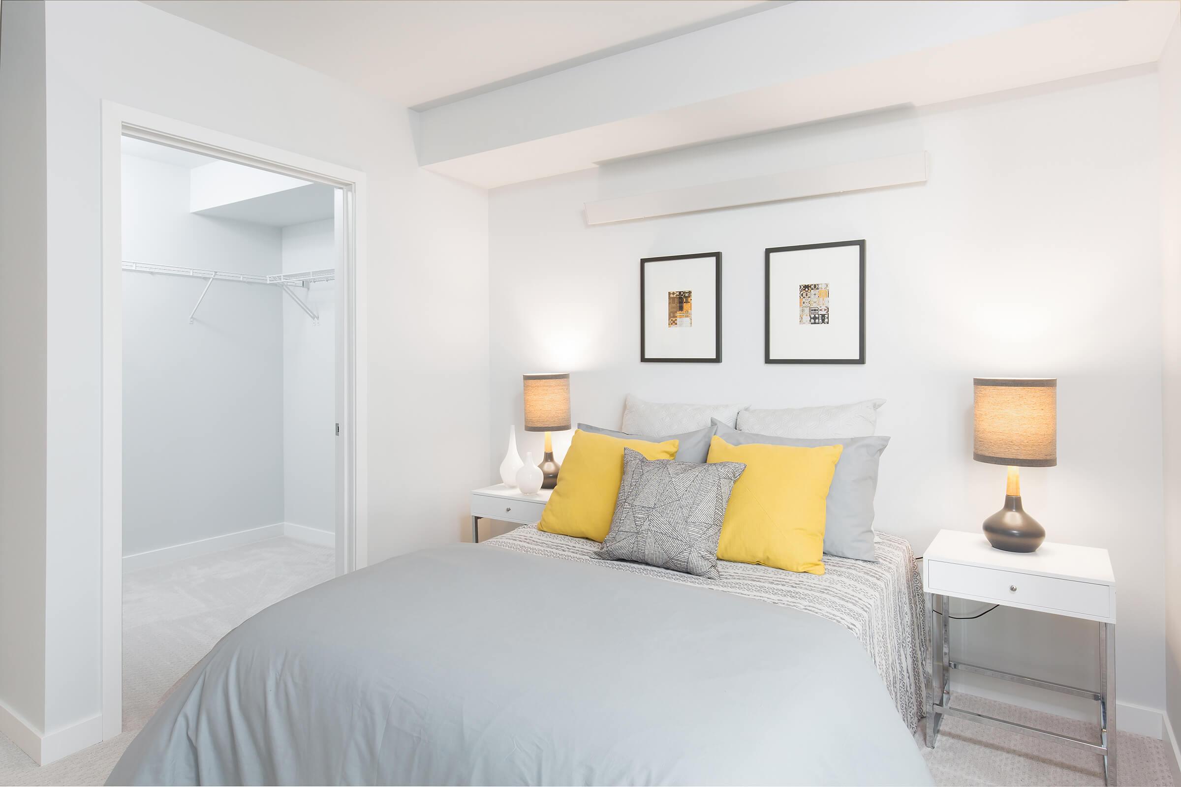 A cozy bedroom featuring a queen-sized bed with gray bedding and yellow accent pillows. Two decorative frames hang above the bed. On either side, there are white nightstands with table lamps. A spacious closet is visible in the background, showcasing a minimalist design and light gray walls.