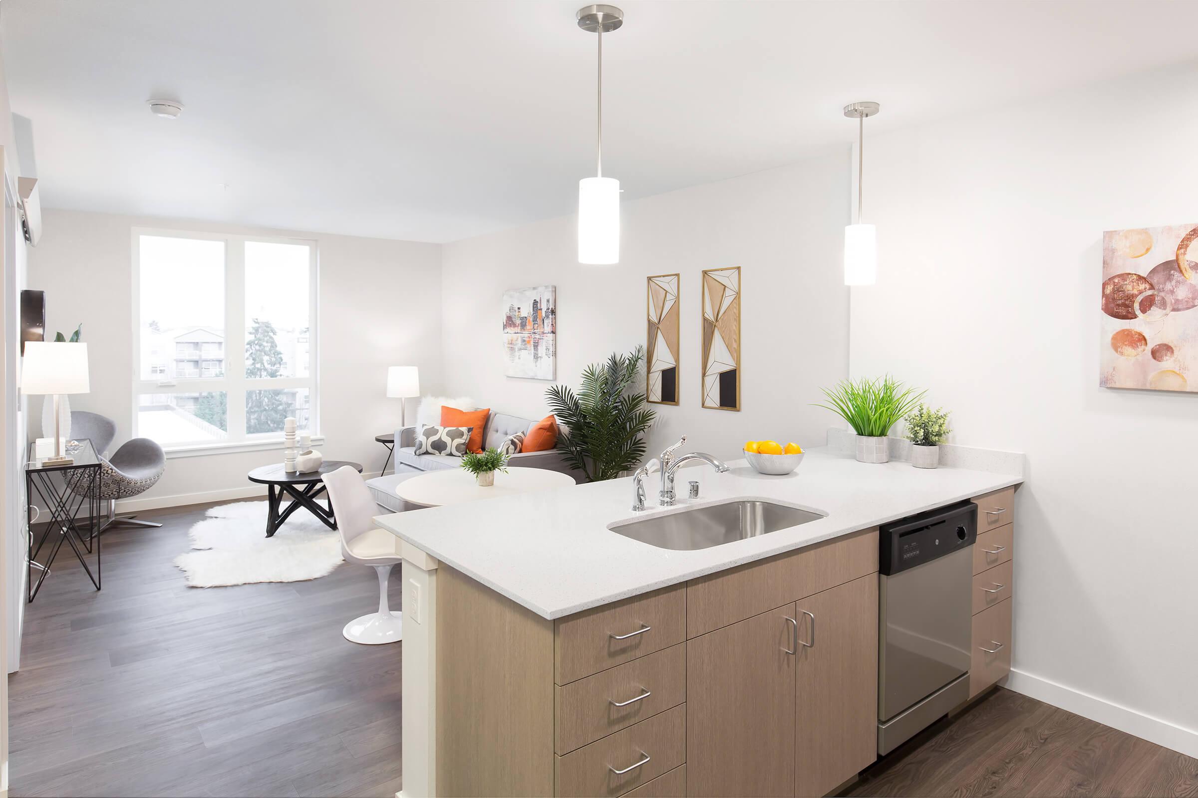 A modern, minimalist kitchen and living area featuring a white countertop with a sink, stainless steel dishwasher, and hanging light fixtures. A cozy living space is visible in the background with a gray armchair, a coffee table, decorative plants, and artwork on the walls. Bright natural light enters through large windows.