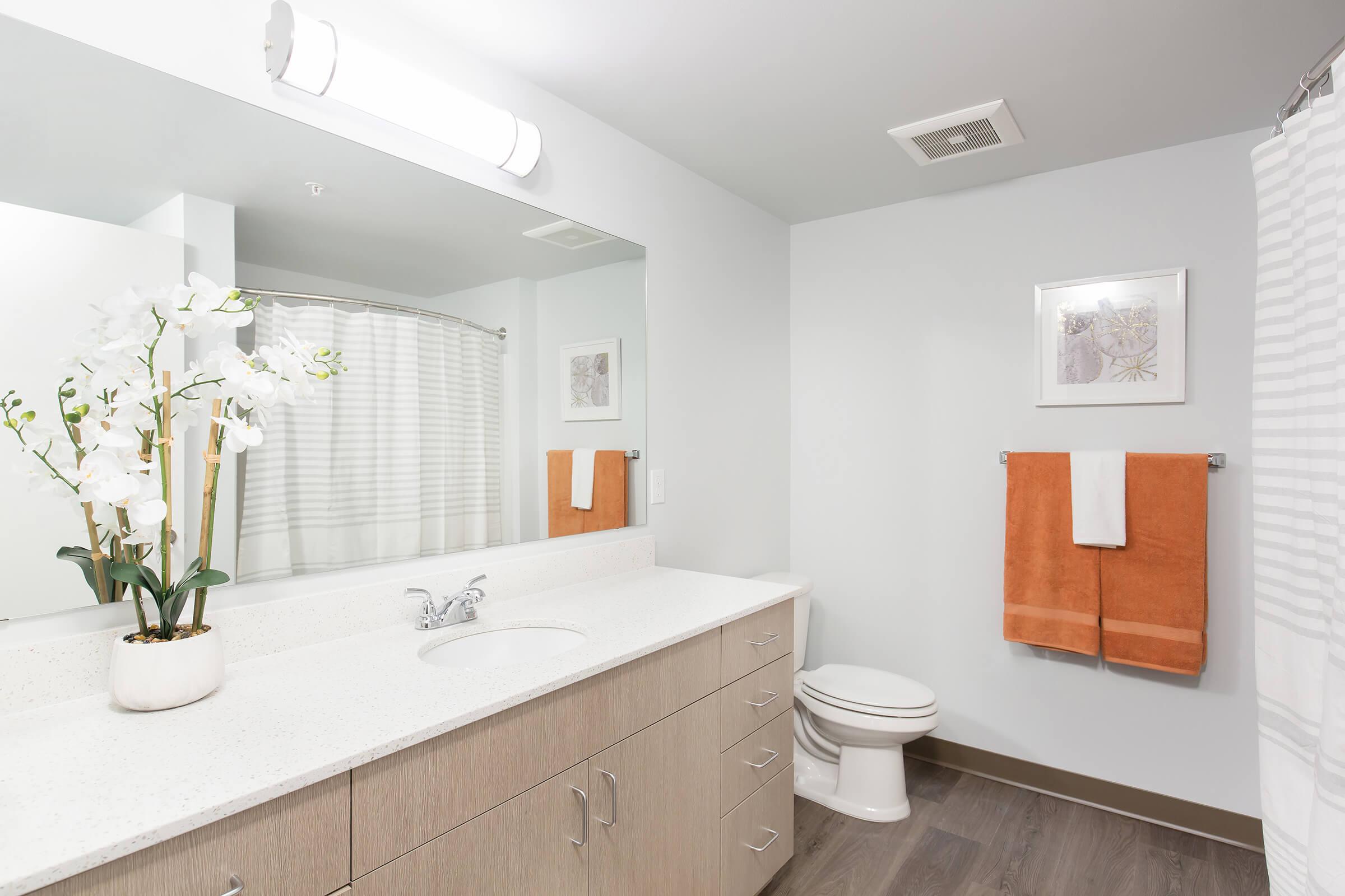 A modern bathroom featuring a light-colored countertop with a sink, a white shower curtain with subtle stripes, and orange towels hanging on a towel rack. A potted orchid adds a decorative touch, while a framed artwork is displayed on the wall above the sink. The flooring is a dark wood finish.