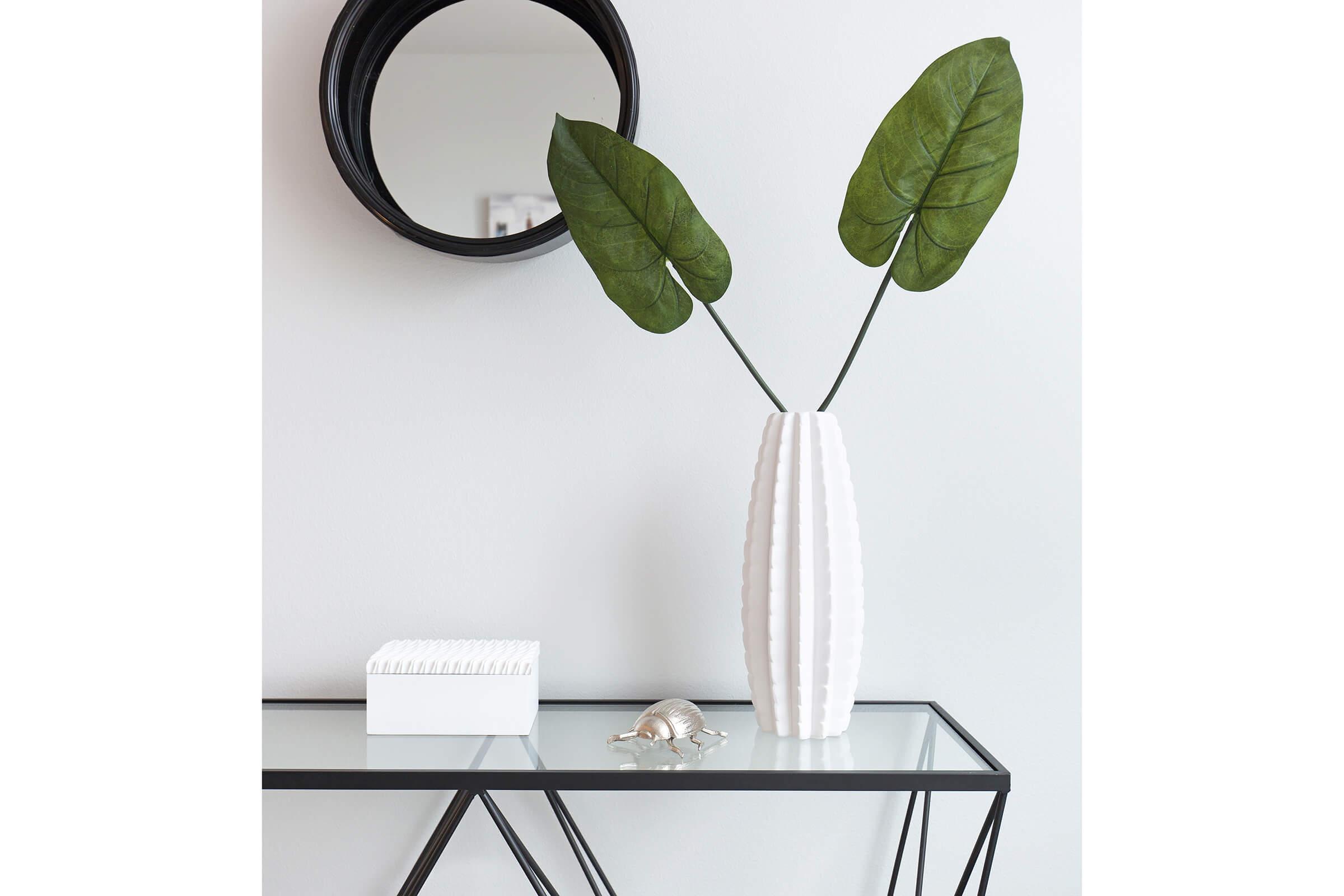 A modern console table featuring a glass top and a geometric black frame. On the table, there is a white textured vase with two large green leaves, a small white box, and a decorative silver object that resembles a snail. A round black mirror is mounted on the wall above the table.