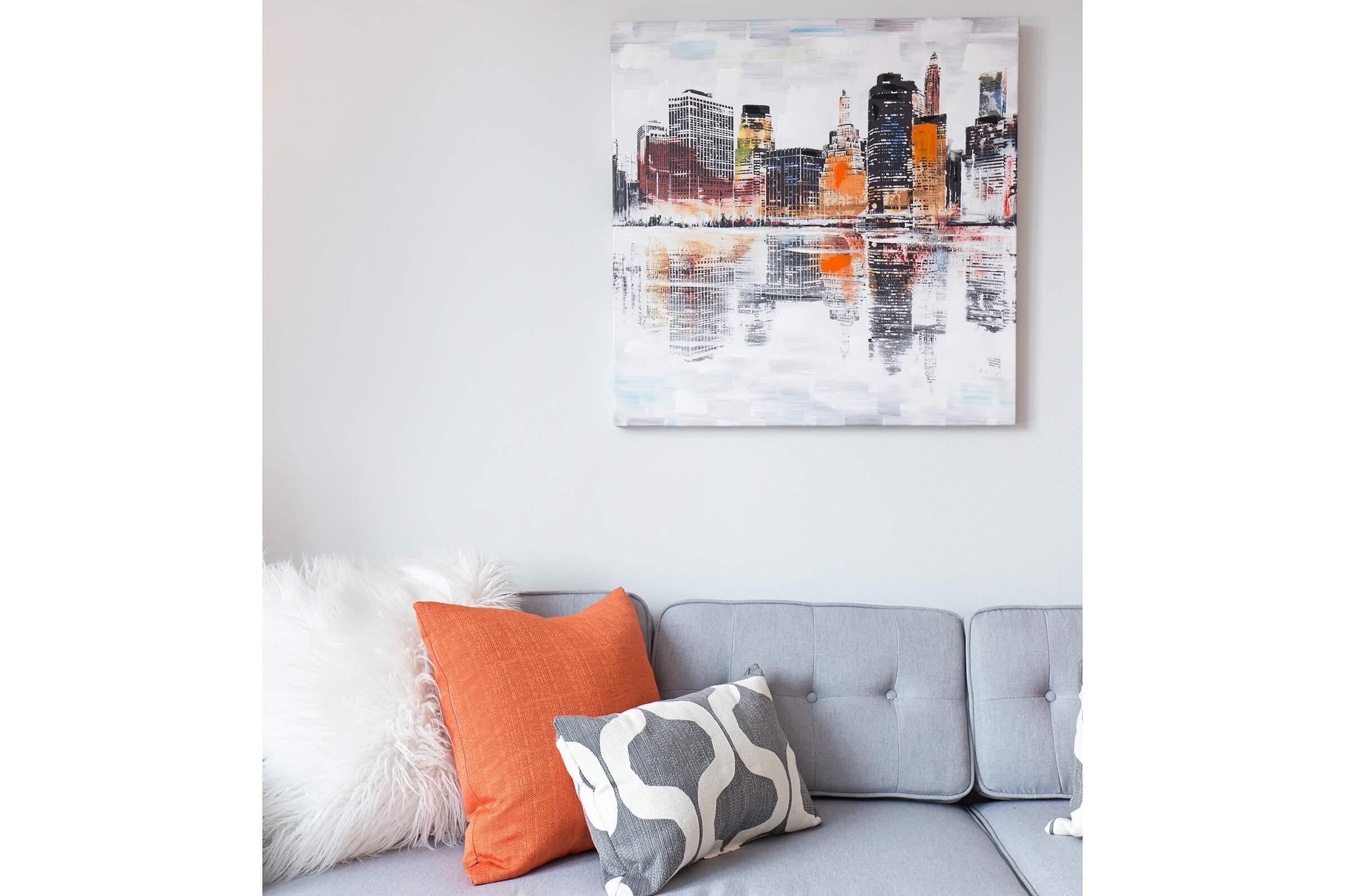 A modern living room scene featuring a gray couch adorned with decorative pillows in orange and patterned gray. Above the couch hangs a colorful abstract painting of a city skyline at dusk, reflecting in water, with warm tones of orange, yellow, and deep blue. A fluffy white throw rests on the couch.