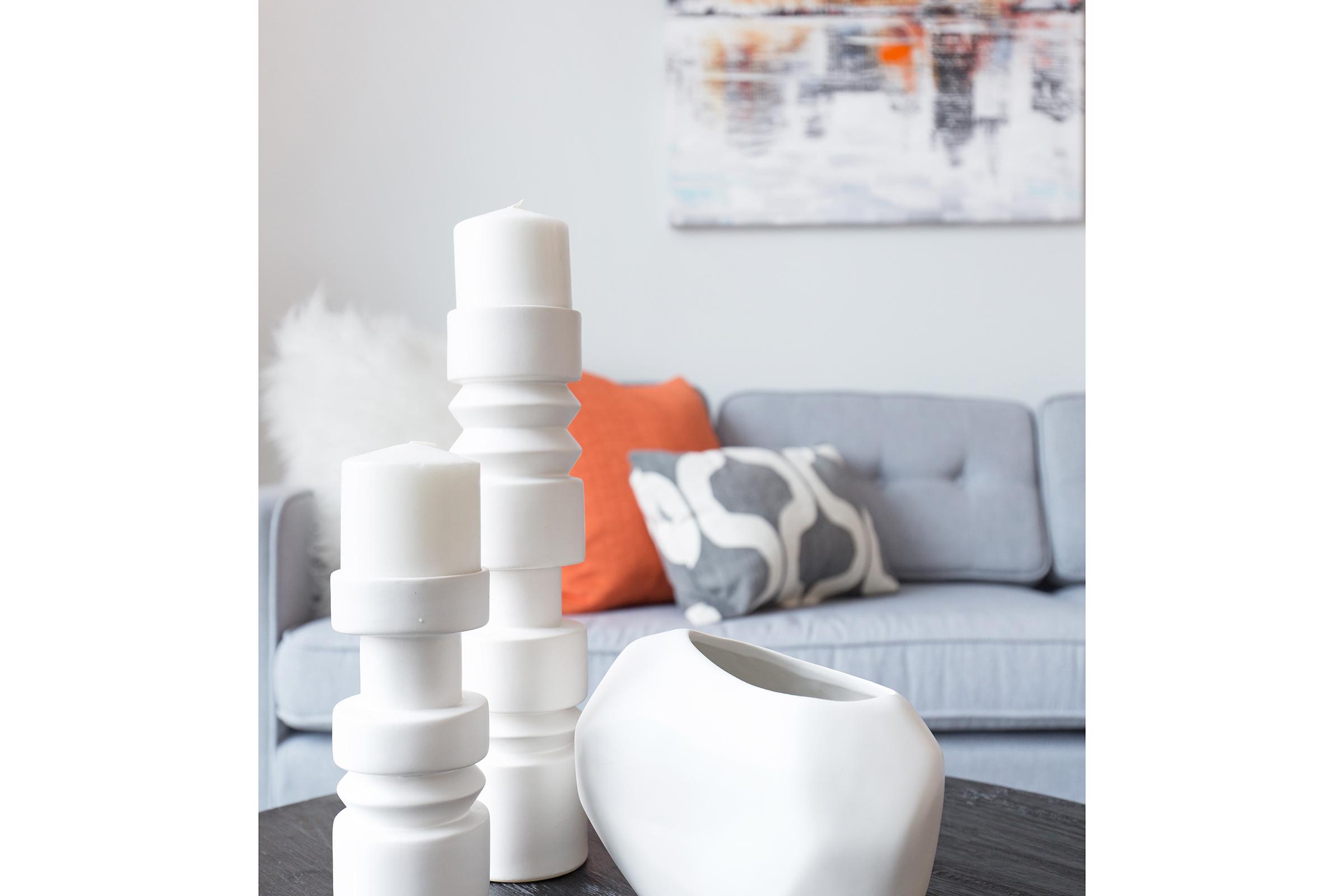 A modern living space featuring a light gray sofa adorned with an orange and gray patterned pillow. In the foreground, there are several white decorative candles of varying heights and a smooth, white vase, creating a stylish and contemporary ambiance. A textured artwork hangs on the wall behind the sofa.