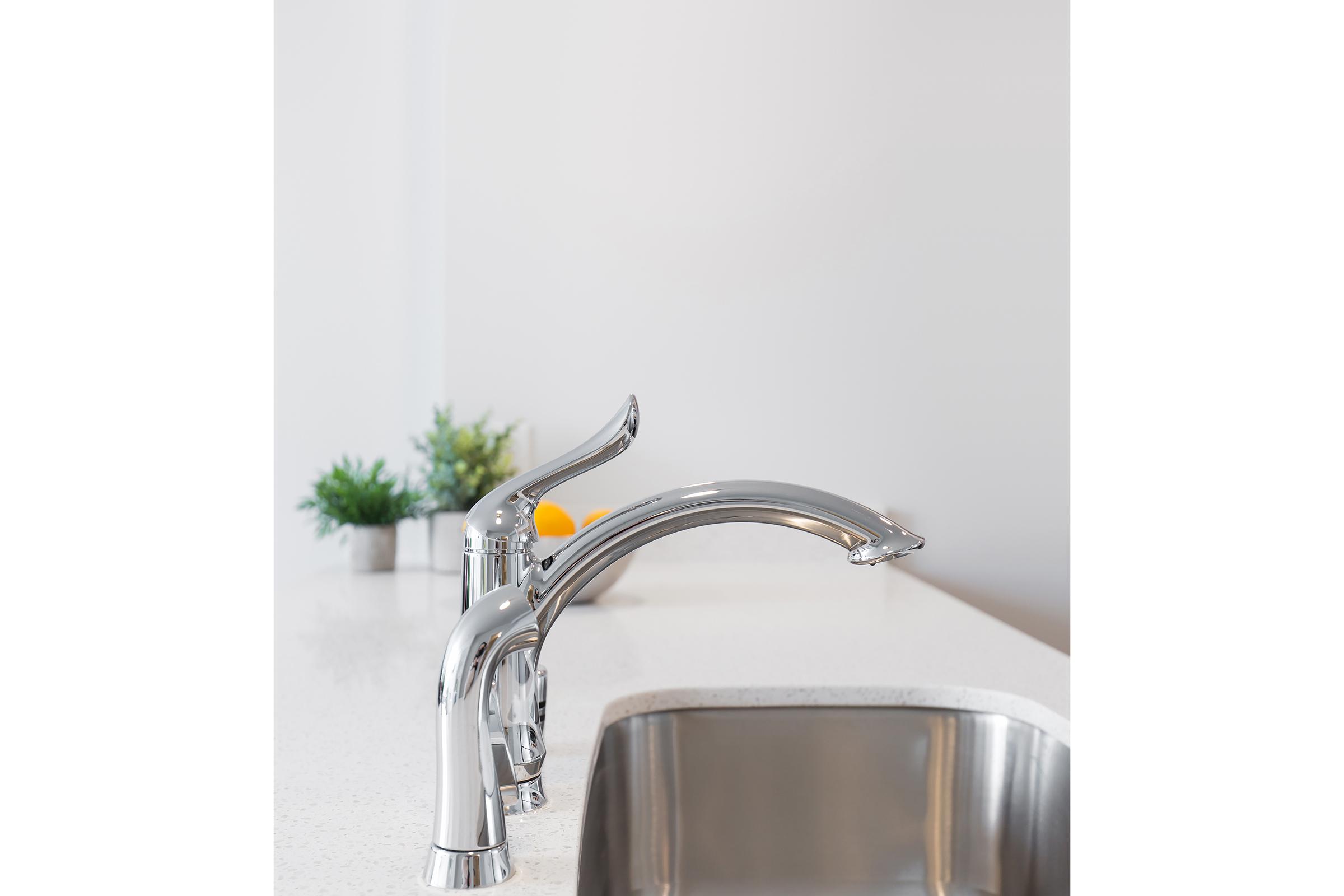 A close-up view of two modern chrome kitchen faucets installed next to a stainless steel sink. The faucets feature sleek, curved designs, and the countertop is light-colored with a simple, minimalistic aesthetic. There is a small potted plant in the background, adding a touch of greenery.