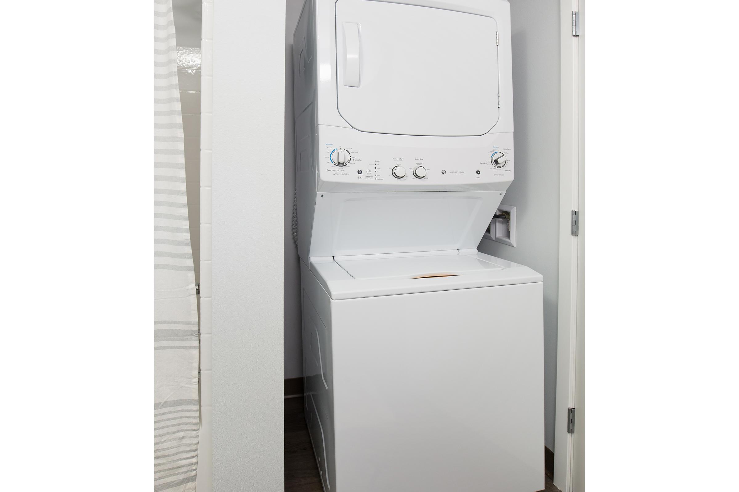 A white stacked washer and dryer unit in a compact laundry space, positioned between walls with light-colored tiles and a curtain in the background. The appliances have visible control knobs and buttons, with the dryer on top and the washer below.