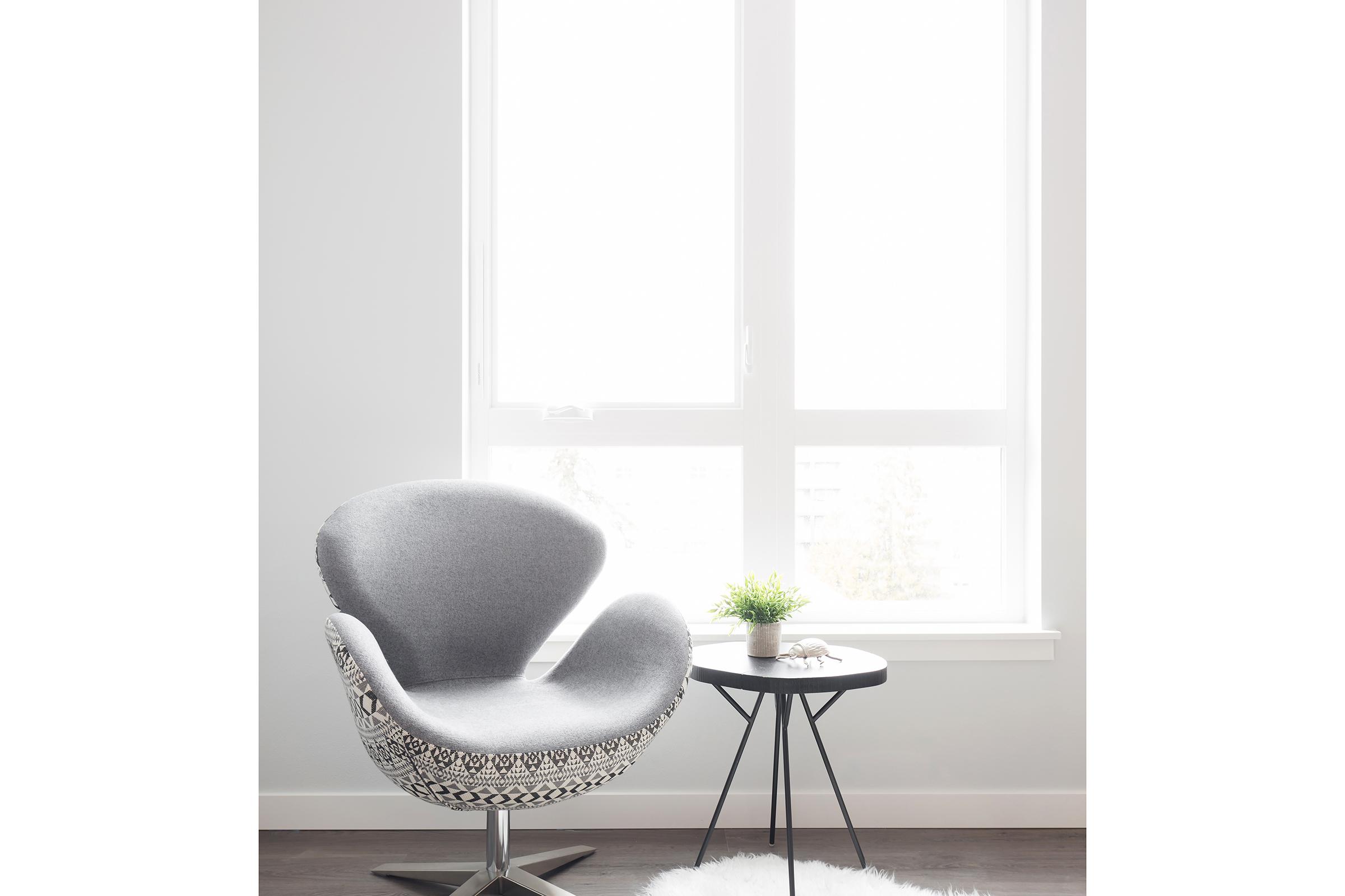 A stylish gray chair with a unique design sits beside a small black side table. The table holds a small green plant and a decorative item. The scene is bright, with light streaming in through large windows, and the floor features a soft white rug, creating a cozy and modern atmosphere.