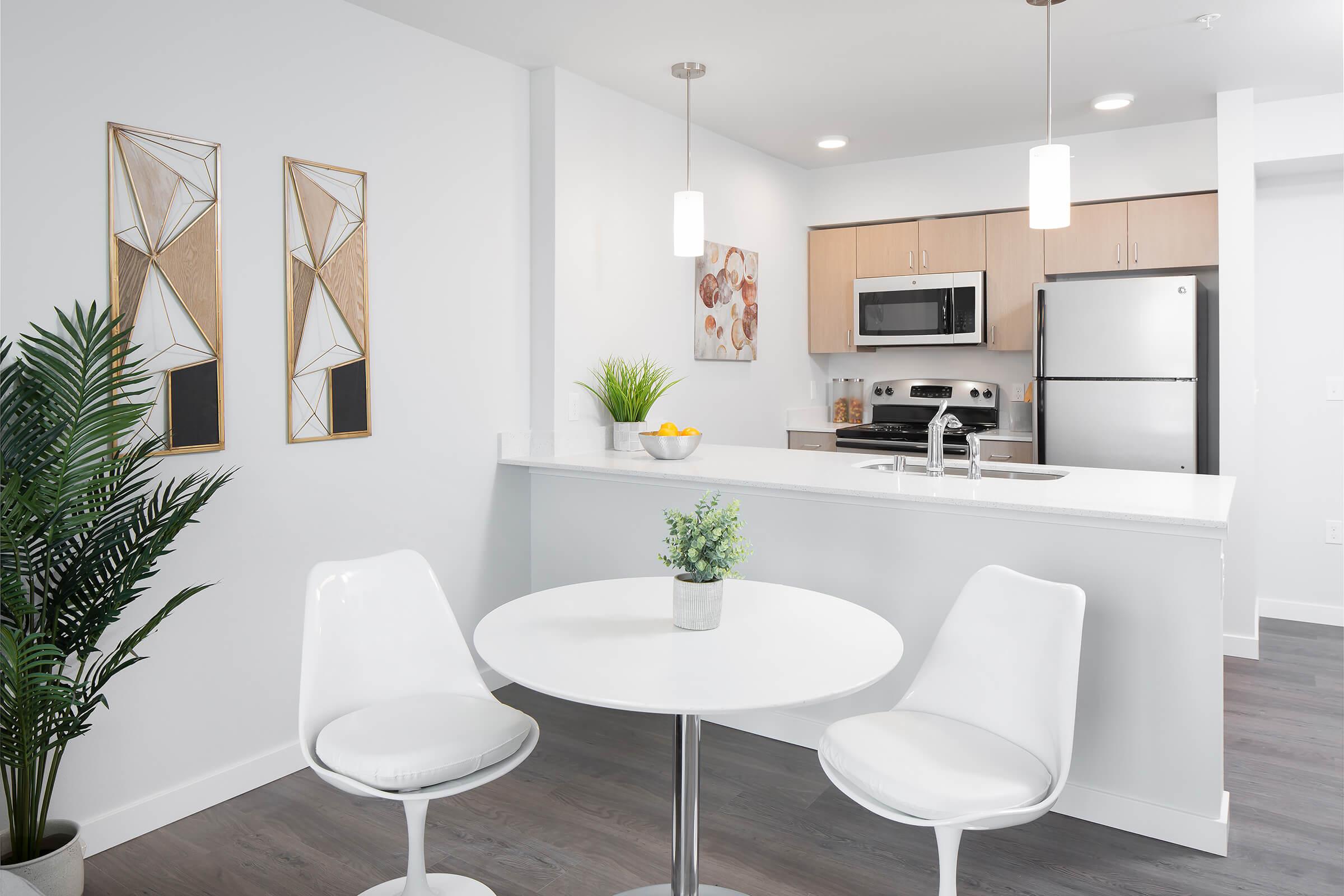 A modern kitchen and dining area featuring a round white table with two white chairs, a decorative plant, and a small bowl of fruit on the table. The kitchen is equipped with stainless steel appliances and light wooden cabinetry, with geometric wall art on the walls. The space is well-lit and has a clean, contemporary design.