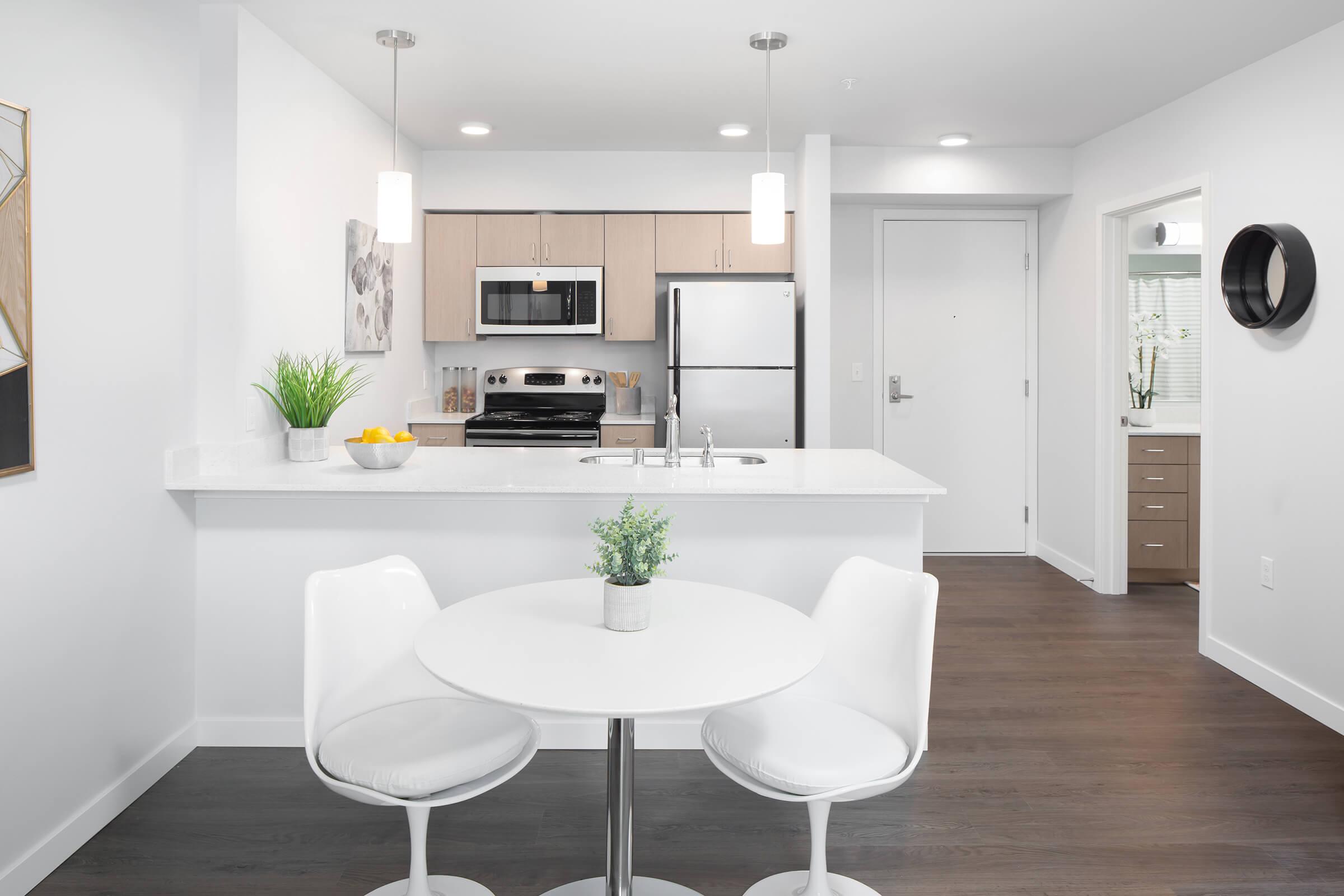 A modern kitchen and dining area featuring a round white table with two white chairs, a stainless steel refrigerator, a stove, and overhead lighting. The kitchen counters are white with a small potted plant and a bowl of yellow fruit. The walls are light-colored, creating a bright and airy atmosphere.