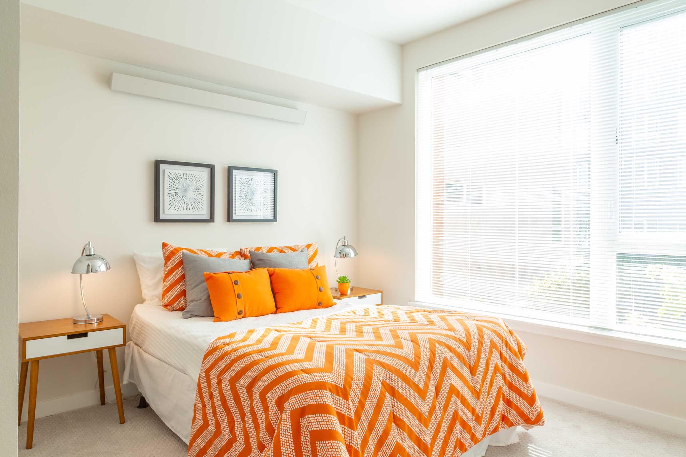 A bright and airy bedroom featuring a bed with an orange and white chevron-patterned blanket and decorative pillows. Two bedside lamps are on either side of a small nightstand. The walls are light-colored, and a large window offers natural light, with sheer blinds allowing a view of the outside.