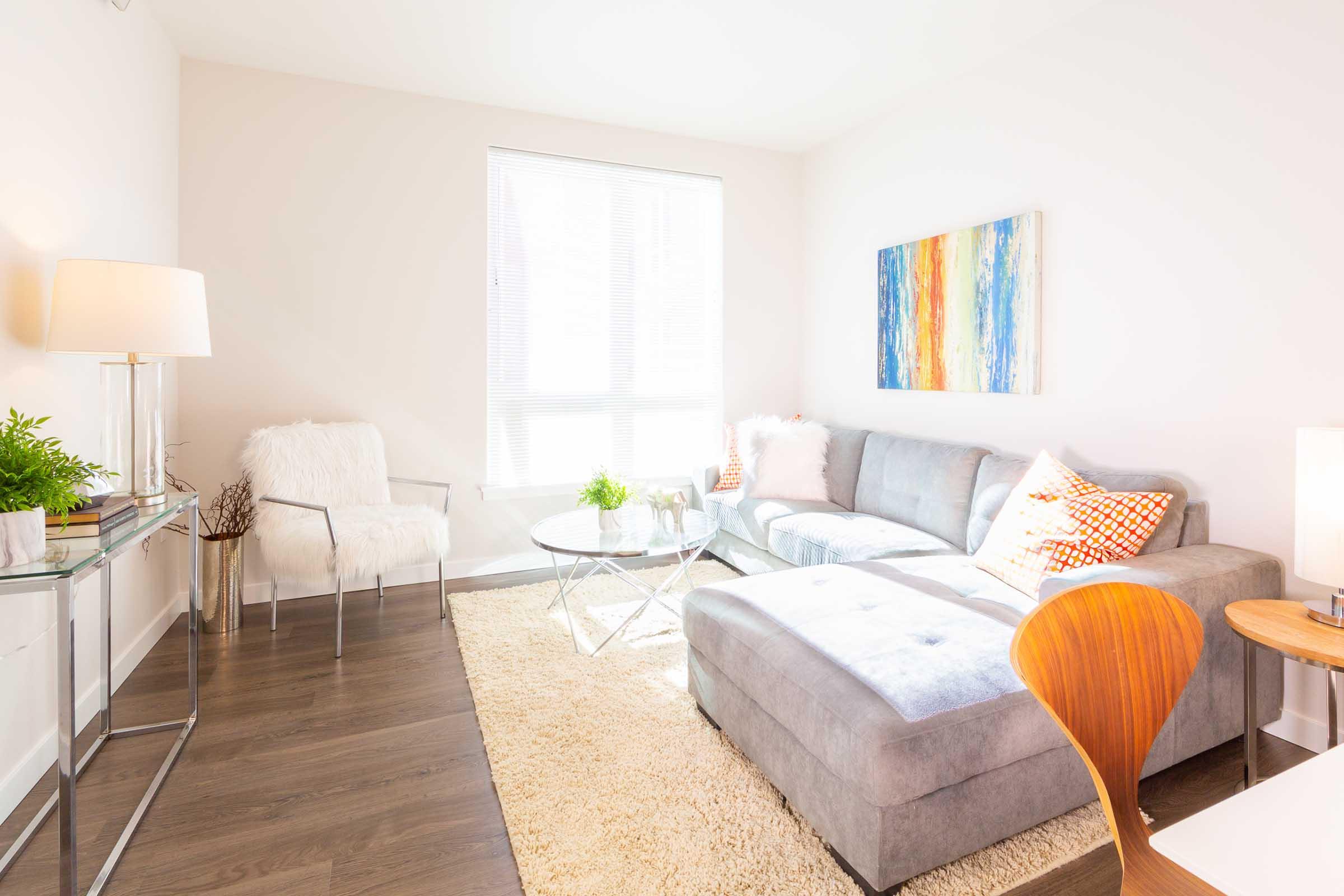 A bright and modern living room featuring a light gray sectional sofa with colorful cushions, a glass coffee table, and a cozy area rug. There are a couple of decorative plants, a stylish chair, and a piece of abstract art on the wall. Large windows allow natural light to fill the space.