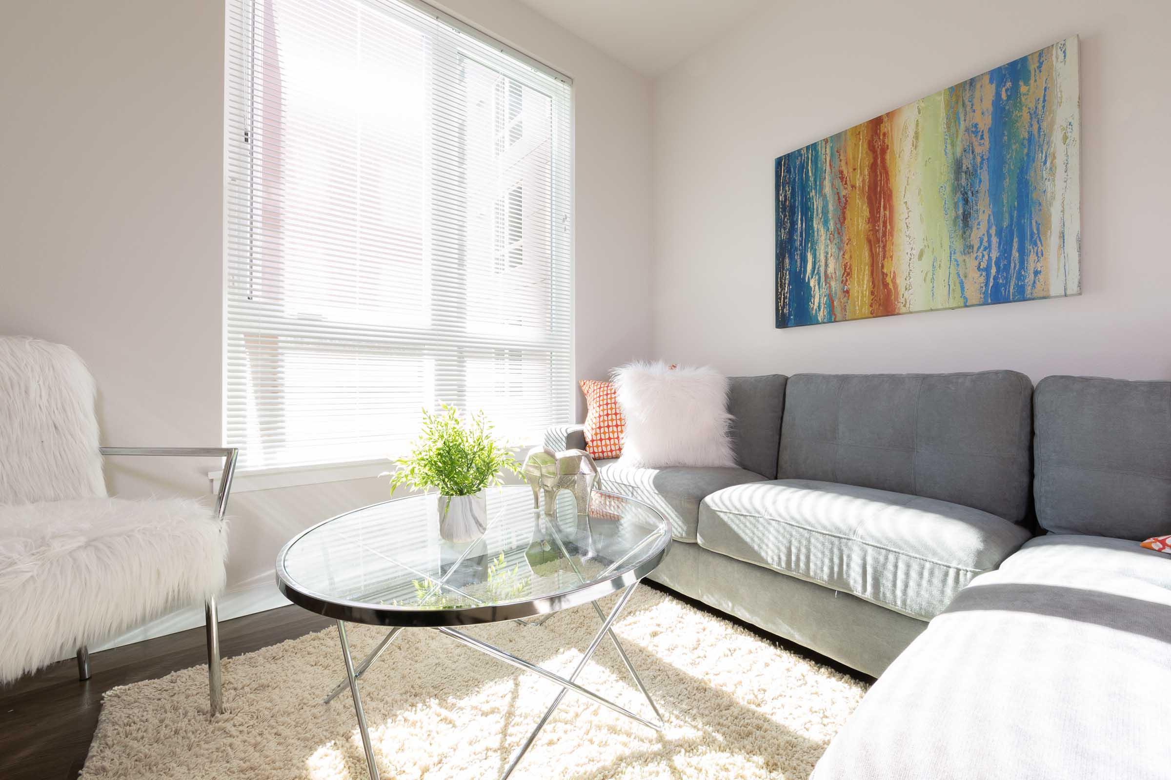 A modern living room featuring a gray sectional sofa, a glass coffee table, and a cozy white chair. Natural light streams through large windows, illuminating a vibrant abstract painting on the wall. A small green plant adds a touch of freshness to the space, complemented by a soft area rug.