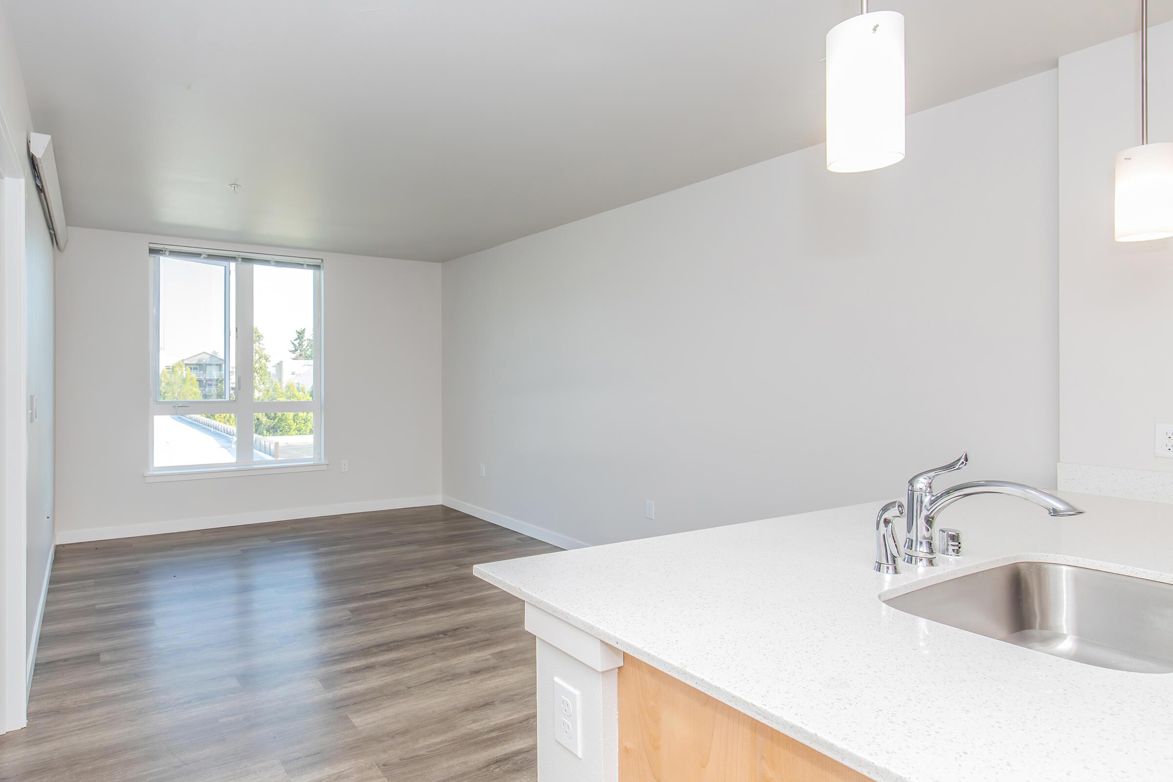 Bright, modern living space featuring a light-colored kitchen counter with a sink, open to a spacious room with large windows. The floor is finished in wood-like laminate, and the walls are painted in a neutral tone. There are two pendant lights above the counter, creating a welcoming atmosphere.