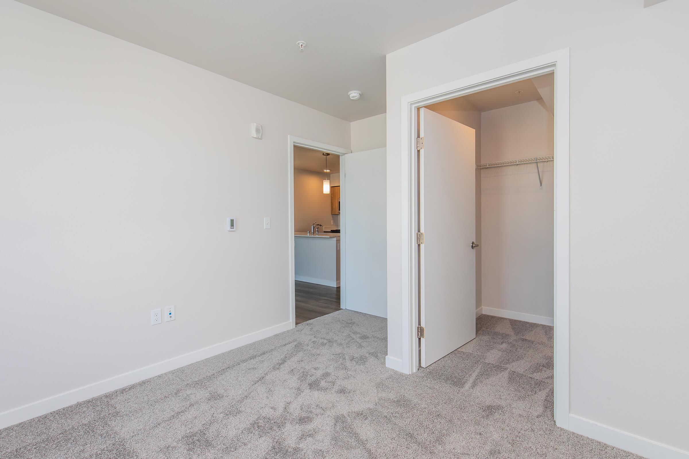 A well-lit, empty room featuring light-colored walls and carpet. On the right, a door leads to a closet. In the background, a glimpse of an adjacent room is visible, with a doorway unoccupied. The space is clean and modern, ideal for personal decoration or furnishing.