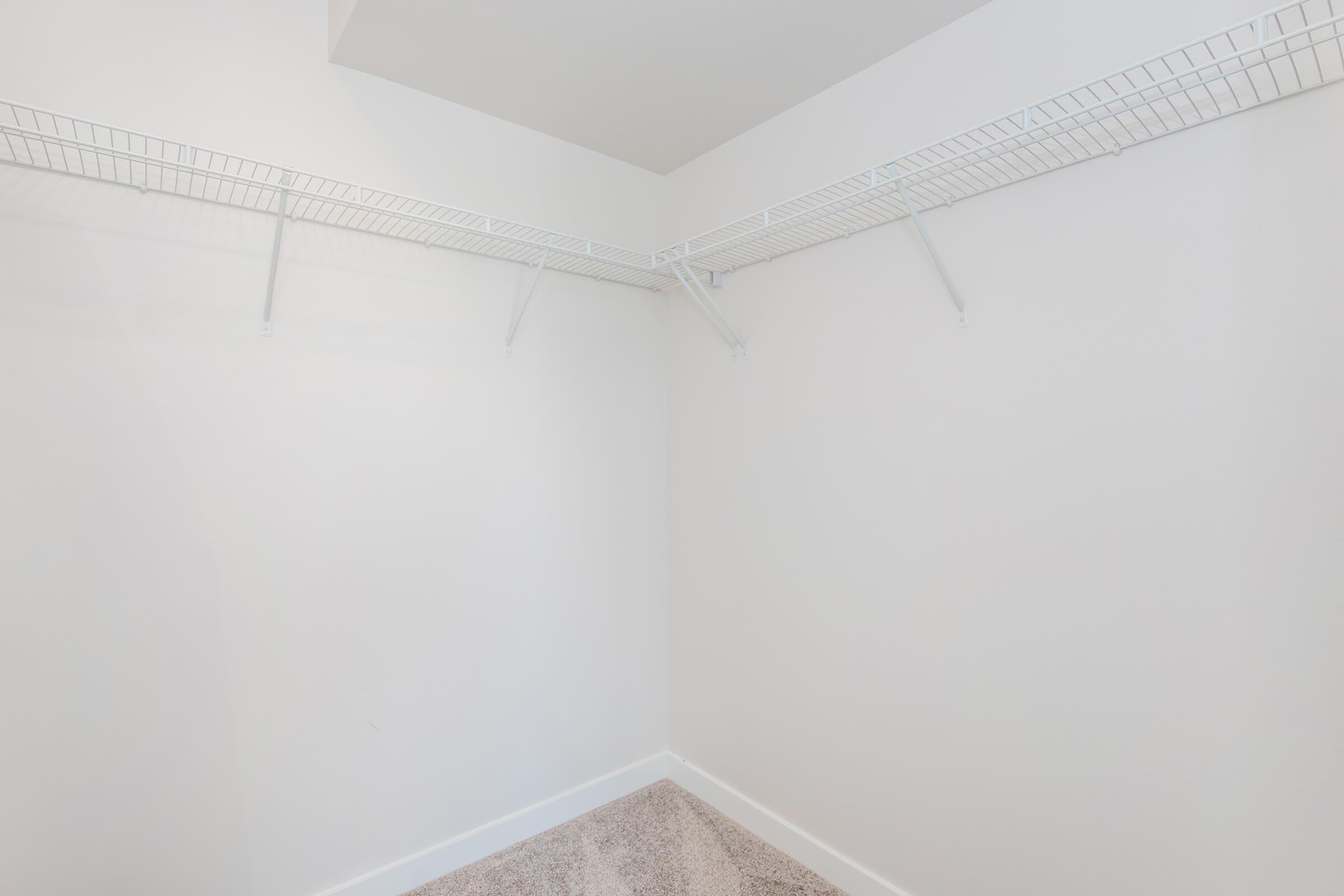 Empty closet with white walls and a light carpeted floor. It features wire shelving along two walls, providing storage space but devoid of any items or decor. The overall appearance is bright and minimalist, emphasizing the spaciousness of the area.