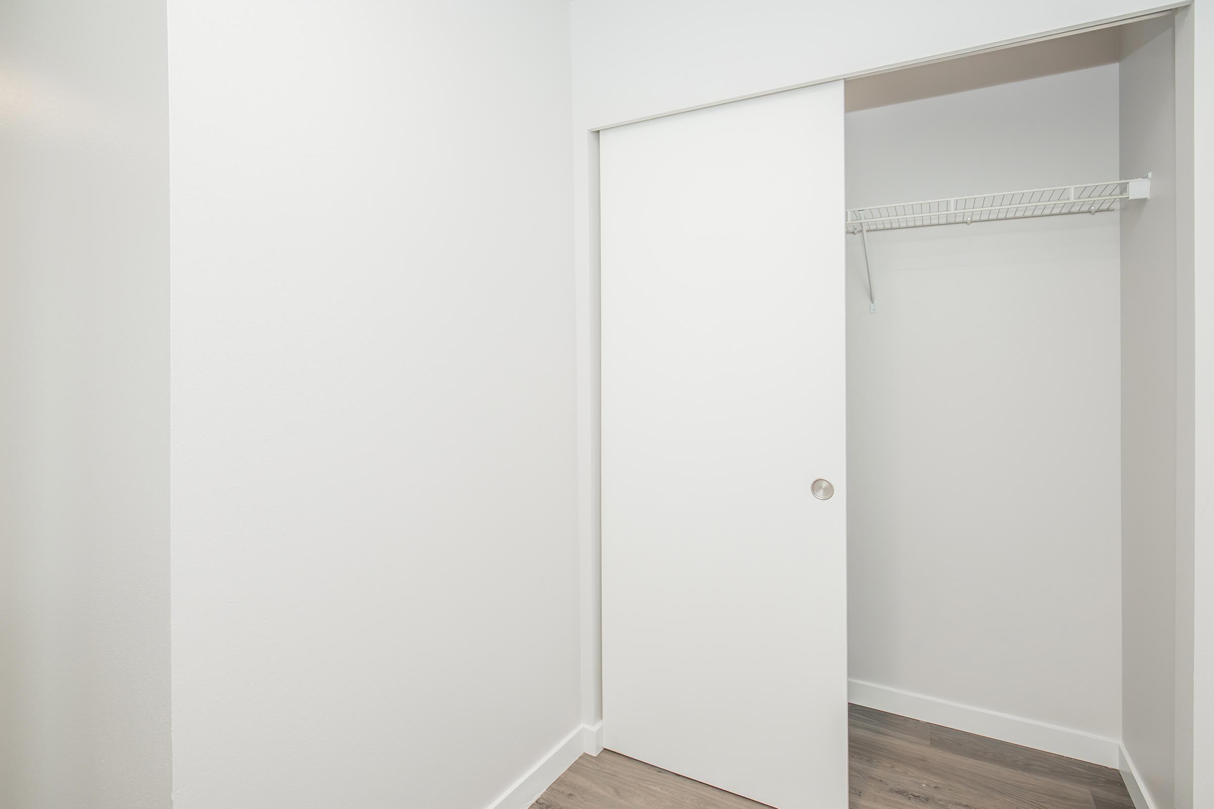 An empty closet with a sliding white door, featuring a built-in wire shelf and plain white walls. The floor has a light wood appearance, giving the space a clean and modern look. The interior of the closet is spacious and uncluttered.