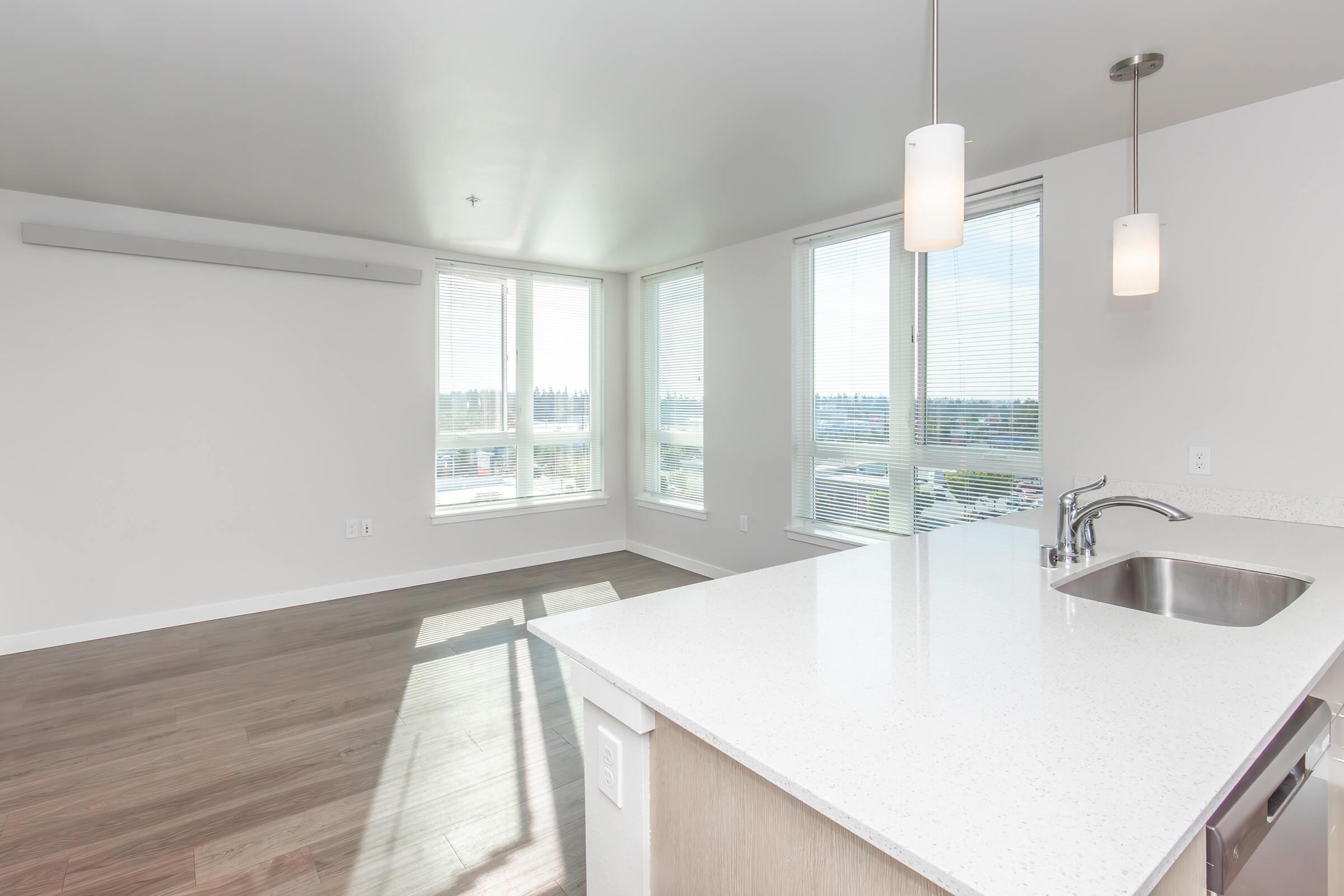 A bright, modern apartment interior featuring large windows with natural light, light-colored walls, and a spacious open layout. The kitchen area includes a countertop with a sink and contemporary pendant lights hanging above. The hardwood floor adds warmth to the space.